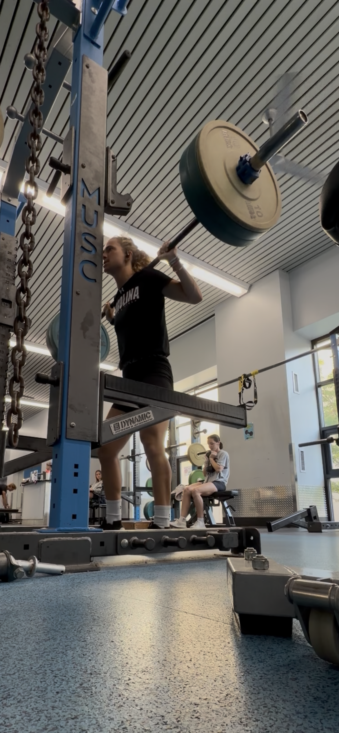 A woman lifting a barbell with weights during a workout in a gym, with another woman sitting on a bench and using her phone in the background.