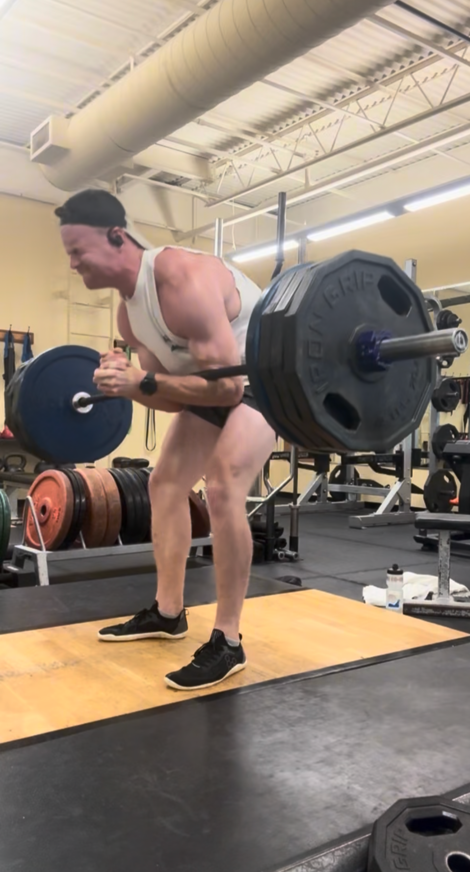 A man lifting a barbell with weighted plates in a gym. He is wearing a tank top, shorts, and sneakers, and appears to be performing a deadlift exercise.