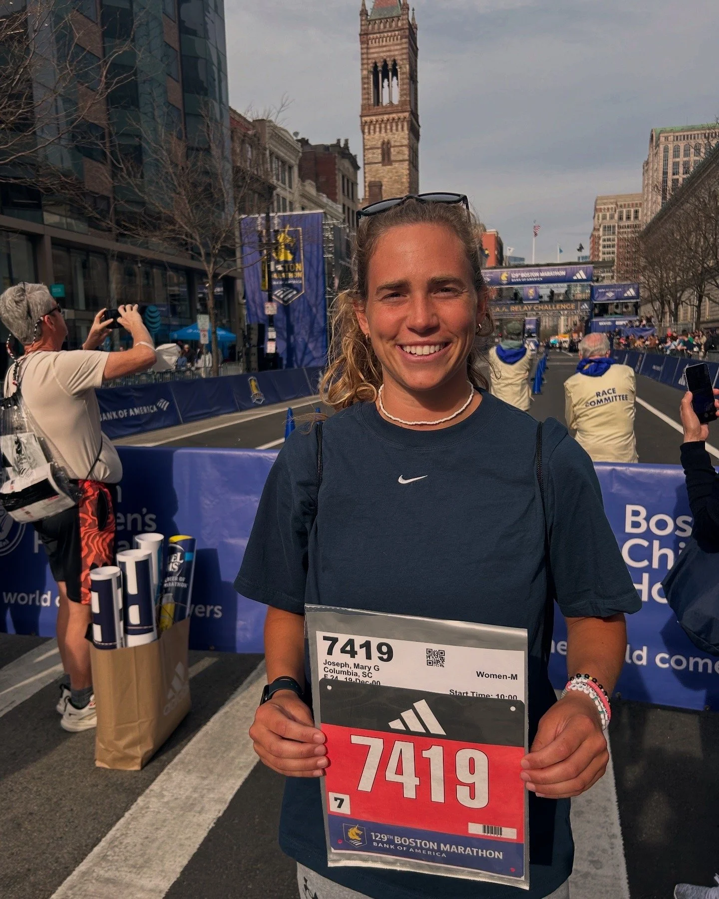 A woman standing at the Boston Marathon starting line, smiling and holding her race bib number 7419. She is wearing a dark T-shirt, a white necklace, and bracelets. In the background, there are other runners, spectators, and city buildings, with a large clock tower visible.