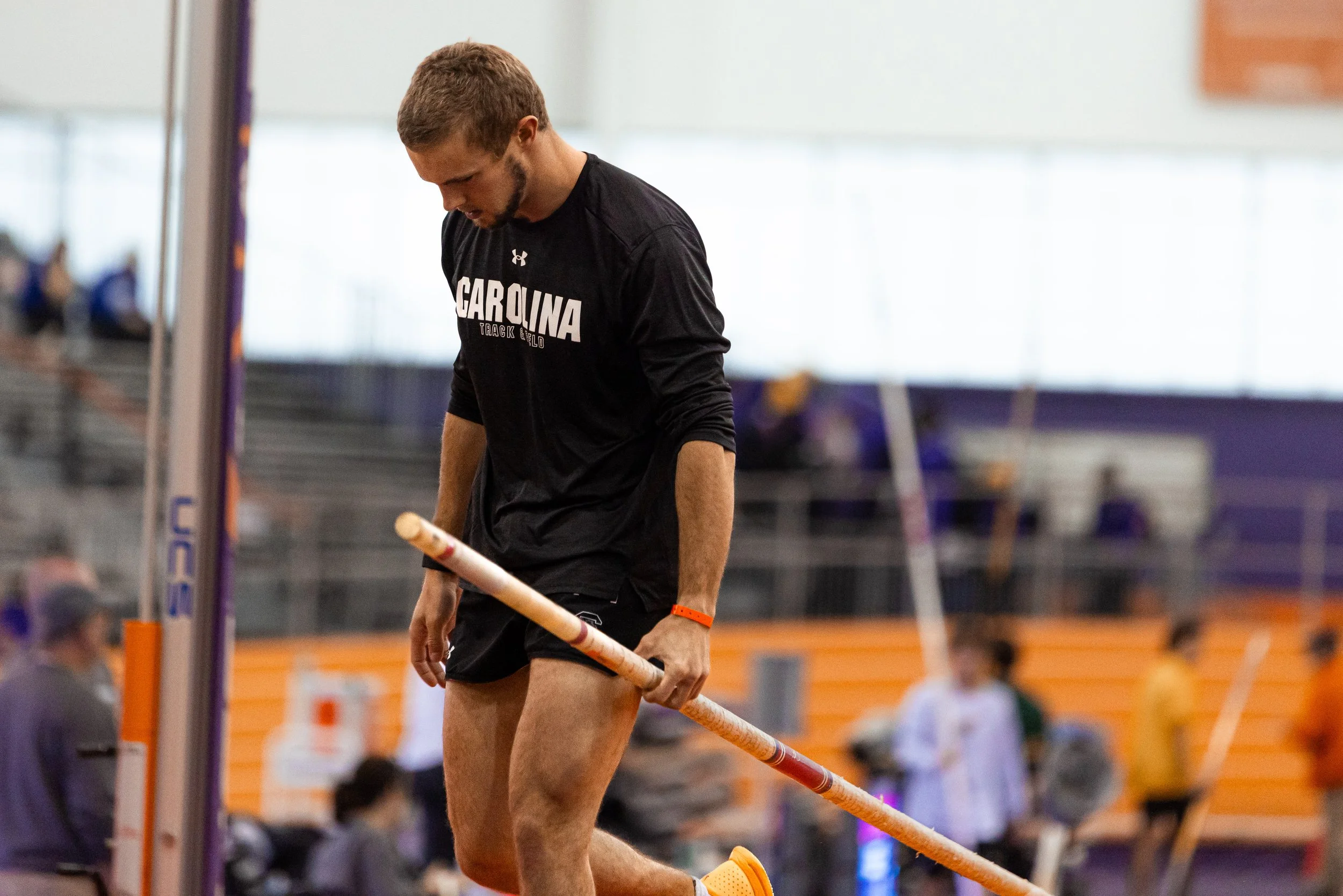 A male athlete in black Carolina Track & Field apparel holding a pole vault pole in an indoor sports facility.