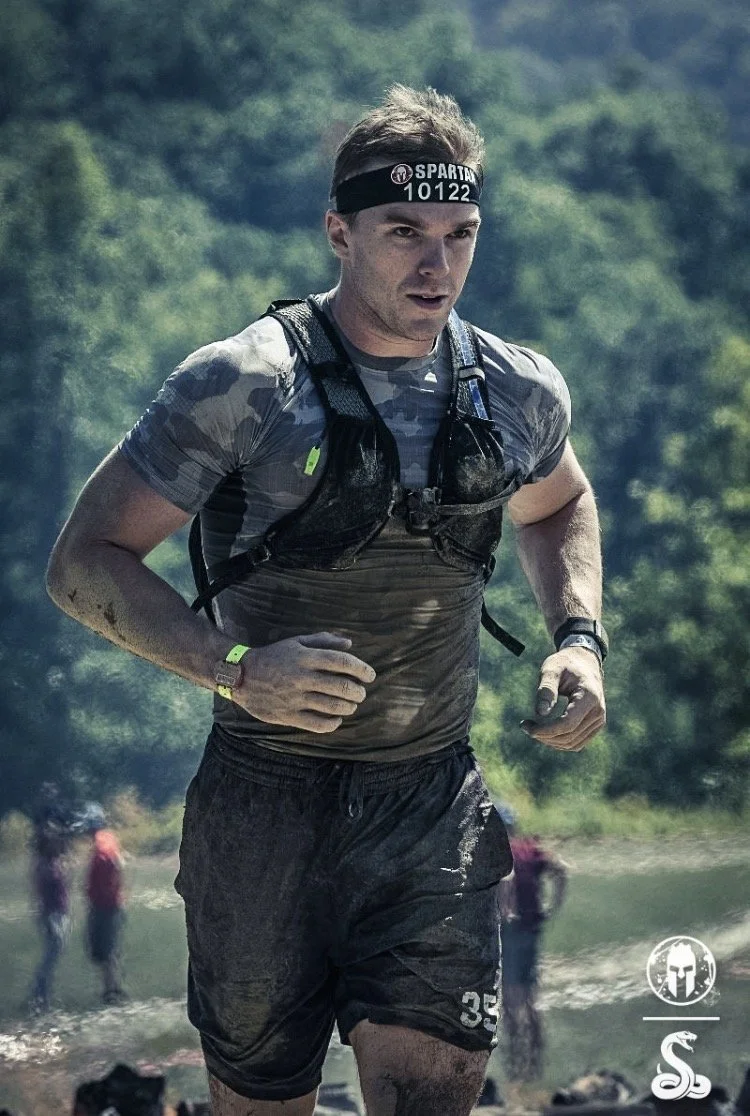 Man participating in an outdoor obstacle course or race, wearing a headband with 'SPARTAN' and number 10122, running through a muddy area with a scenic green forest background.
