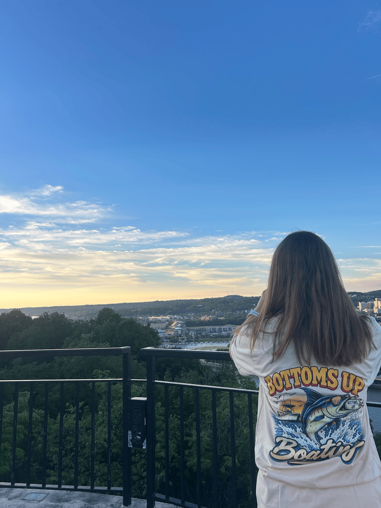 A woman with long brown hair, wearing a white T-shirt with colorful graphics and text, stands on a balcony overlooking a cityscape with trees, buildings, and a hill in the distance under a partly cloudy sky at sunset.