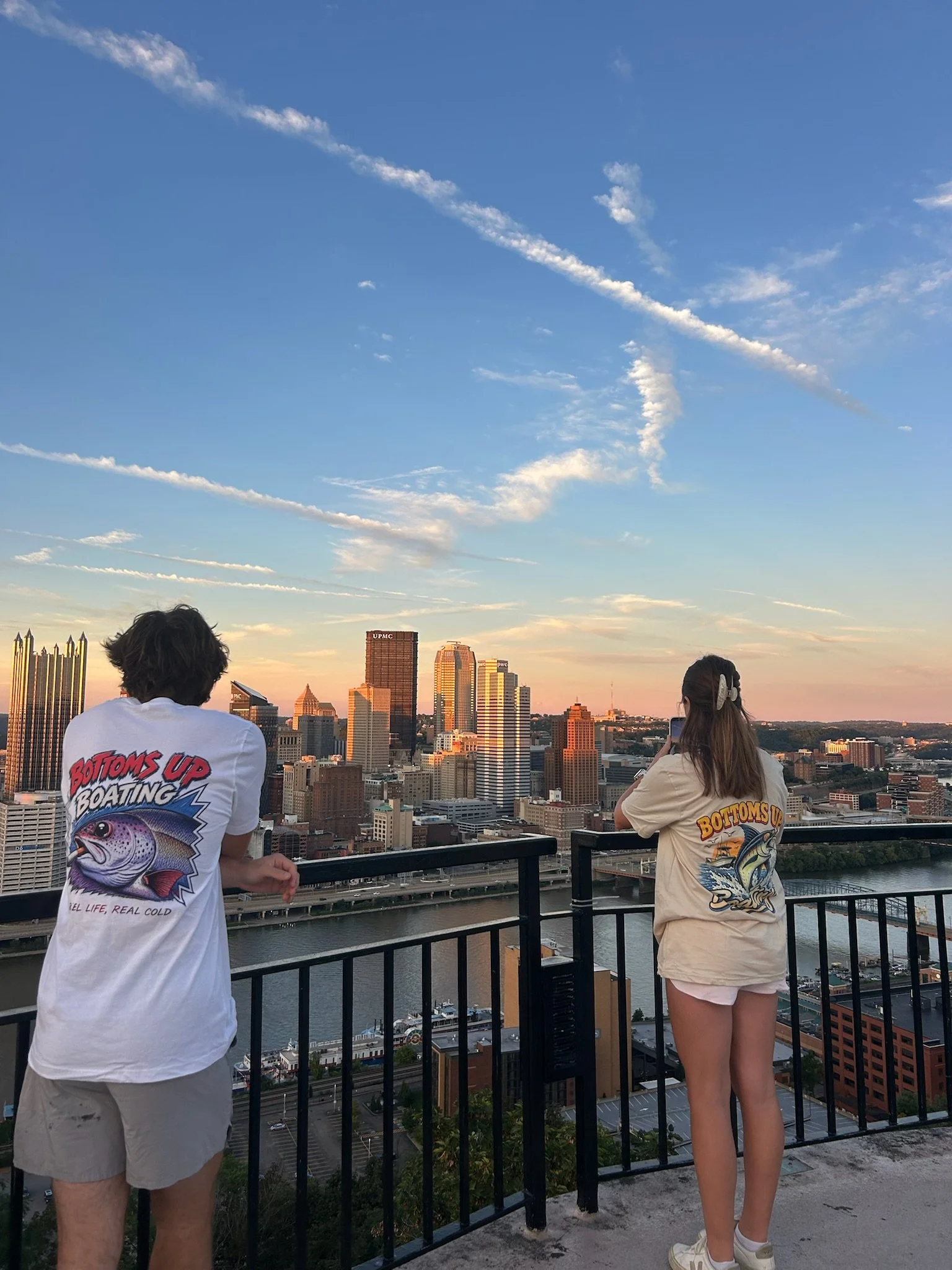 Two people standing on a rooftop balcony looking at a city skyline with tall buildings and a river below during sunset. The person on the left is wearing a white T-shirt with a fish graphic and beige shorts. The person on the right is wearing a beige oversized T-shirt with a fish graphic and white sneakers, holding a phone. The sky has scattered clouds and contrails.