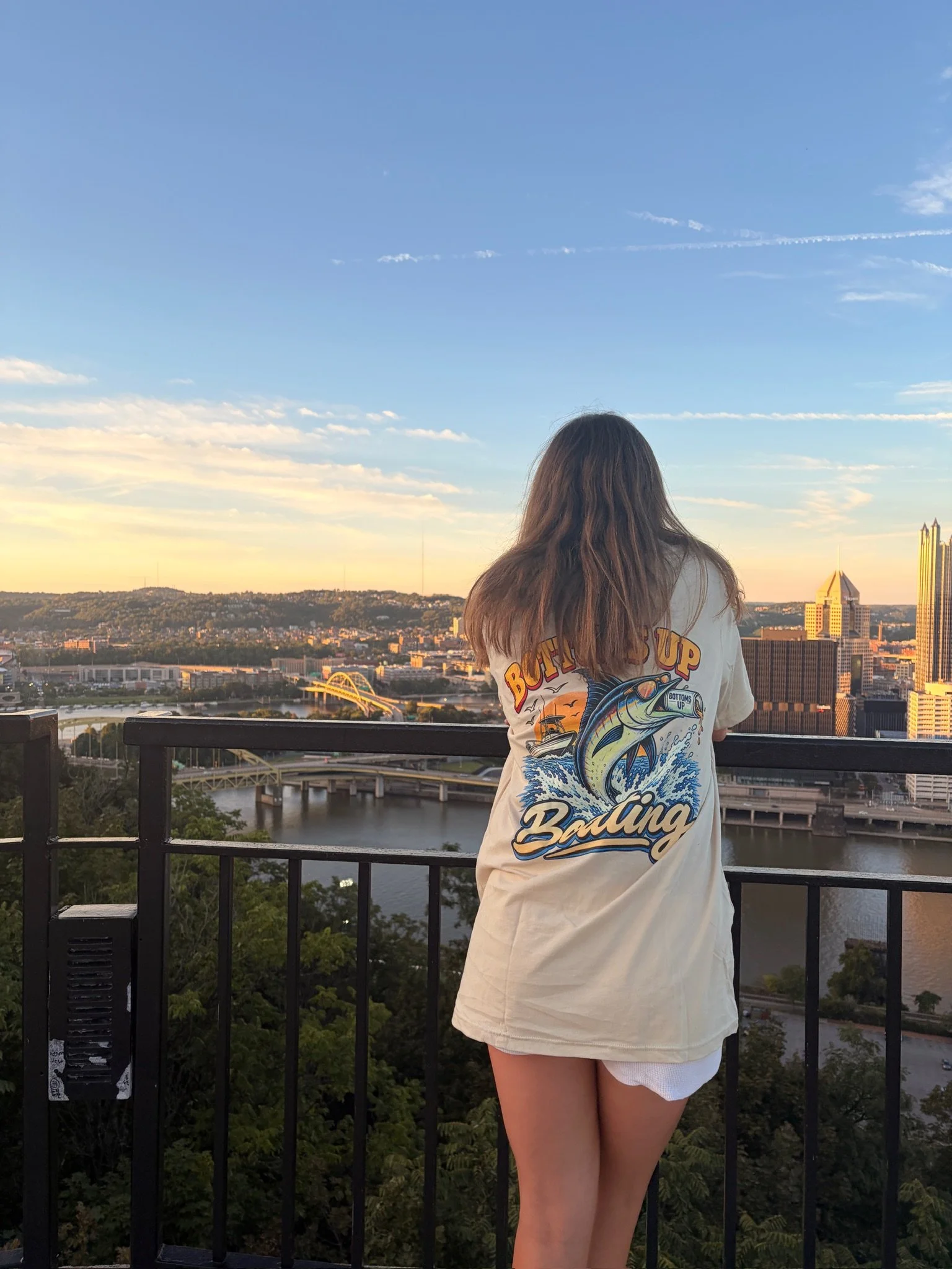 A girl with long brown hair wearing a white t-shirt with fisherman graphics and text, standing on a balcony overlooking a cityscape at sunset, with buildings, a river, and a bridge visible in the background.