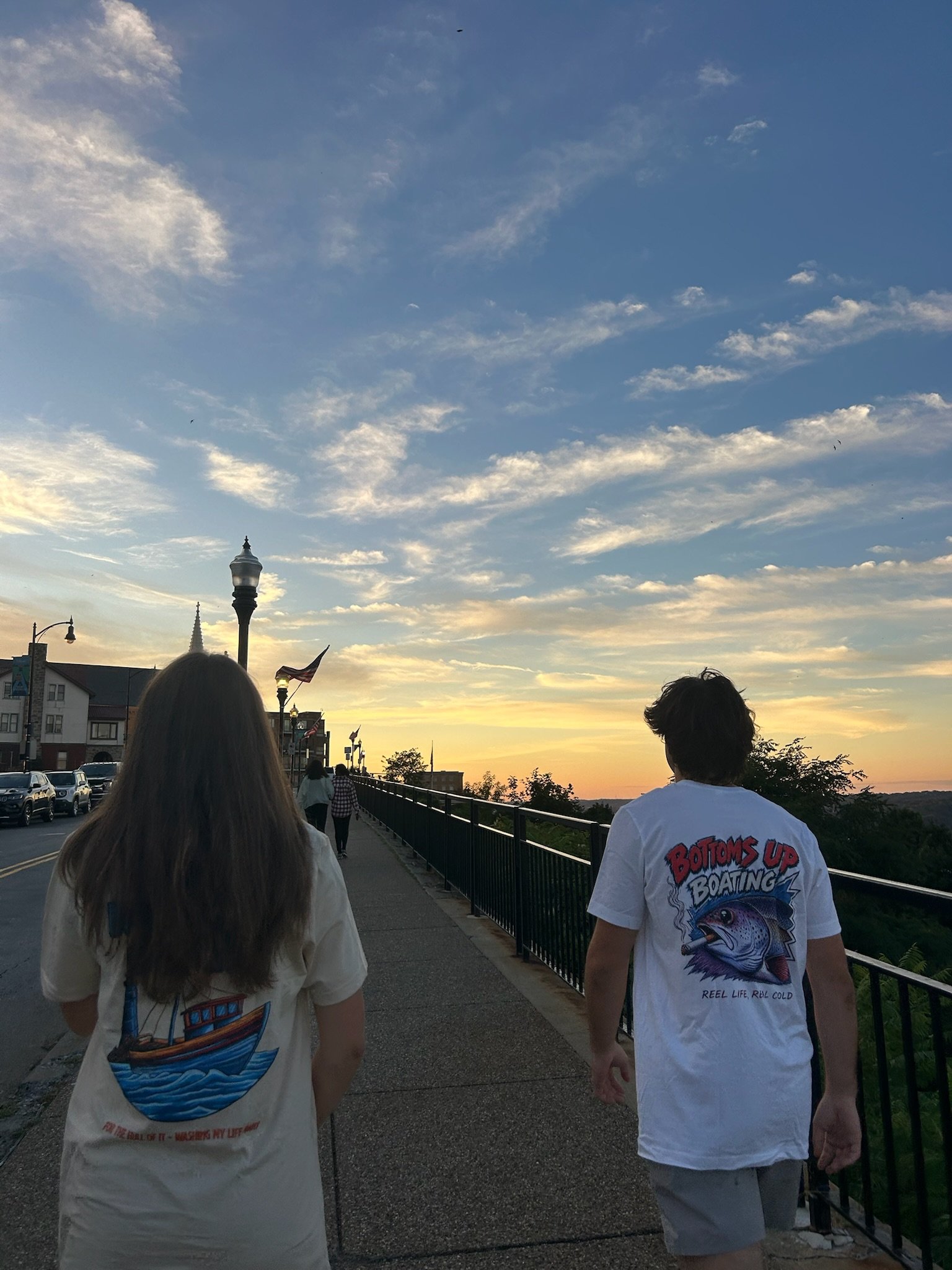 People walking on a sidewalk during sunset, with a cloudy sky, street lamps, and parked cars visible in the background.