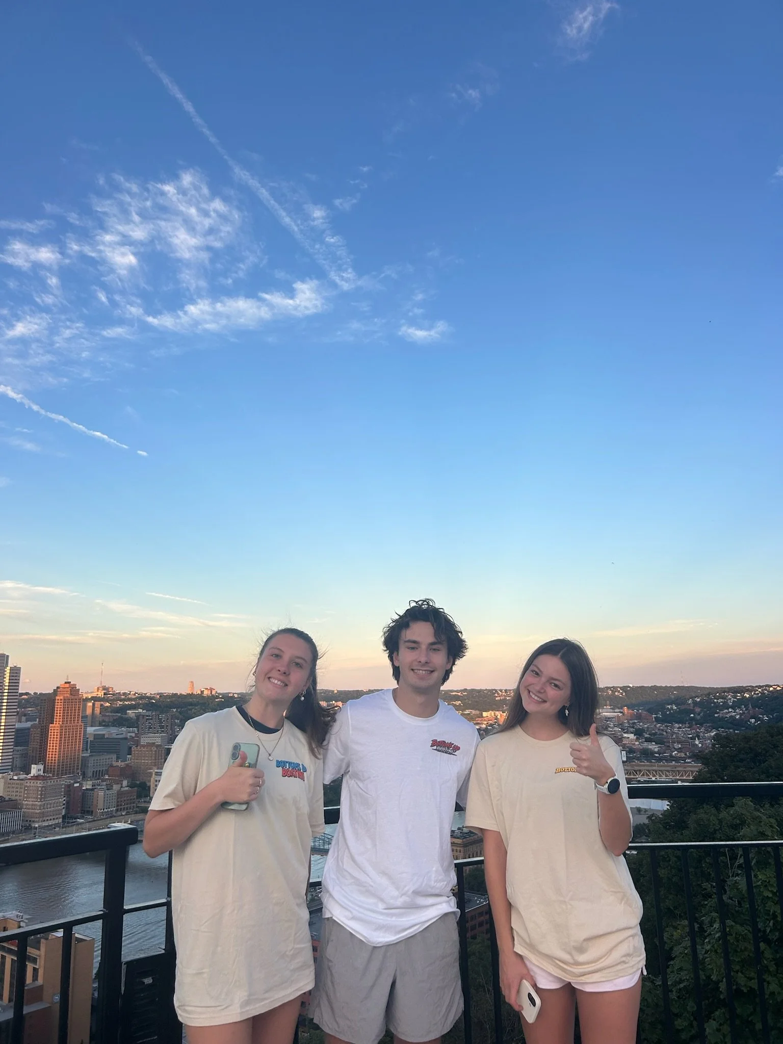 Three young people standing on a rooftop balcony with a city skyline and river in the background during sunset.