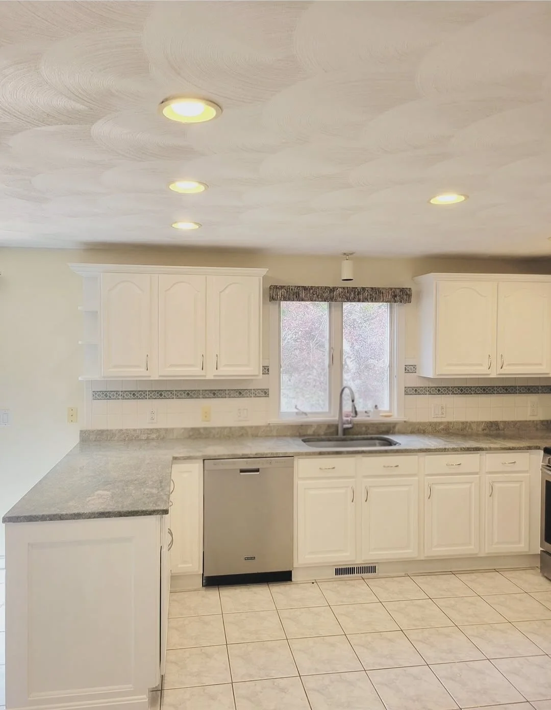 A clean, white kitchen with white cabinets, a granite countertop, a stainless steel dishwasher, a window above the sink, and a tiled floor.