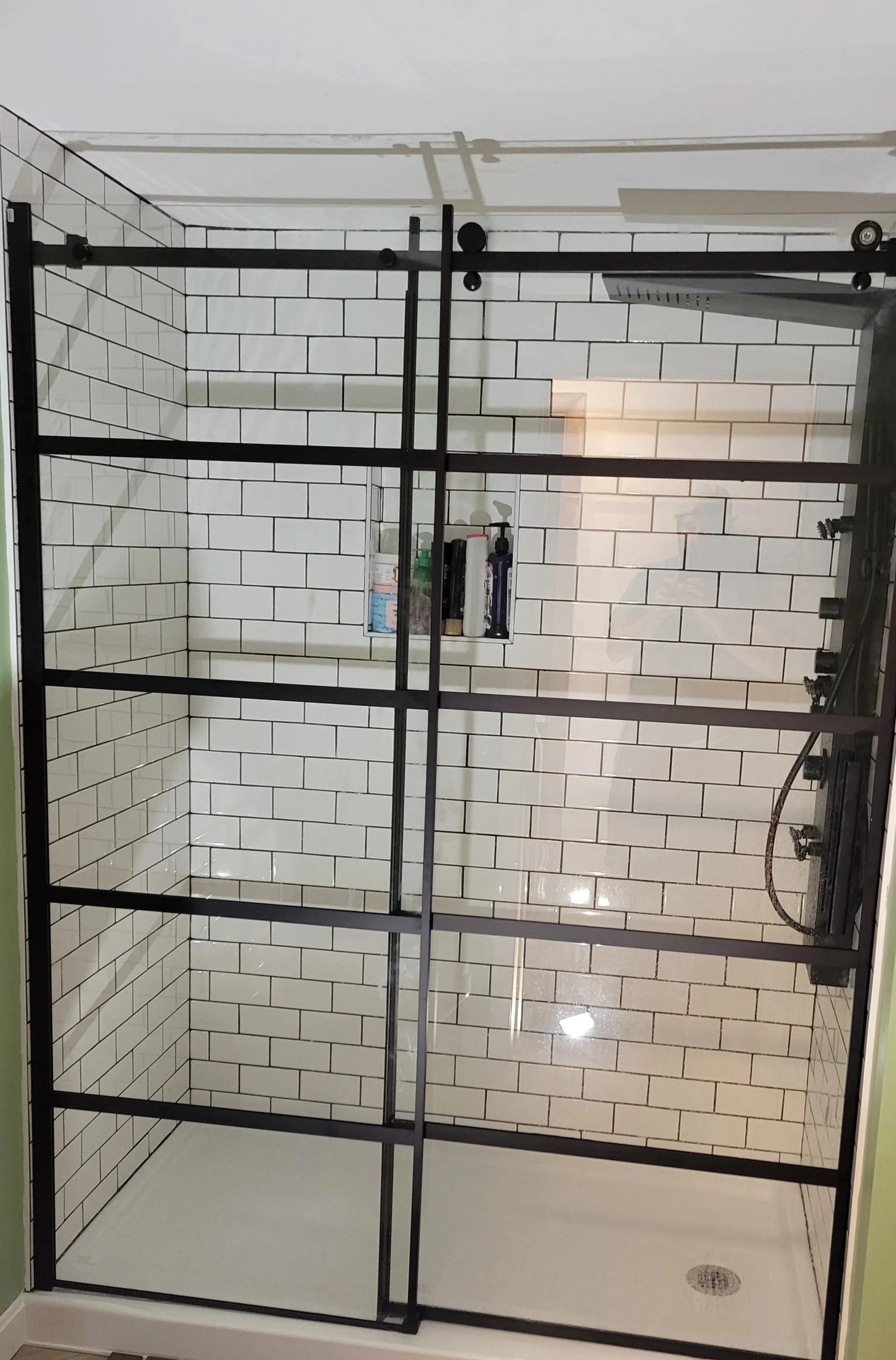 Modern glass shower with black metal frame, white subway tiles on the walls, and a built-in shelf with toiletries. The shower has a rain shower head and a handheld shower head, with a drain on the white floor.