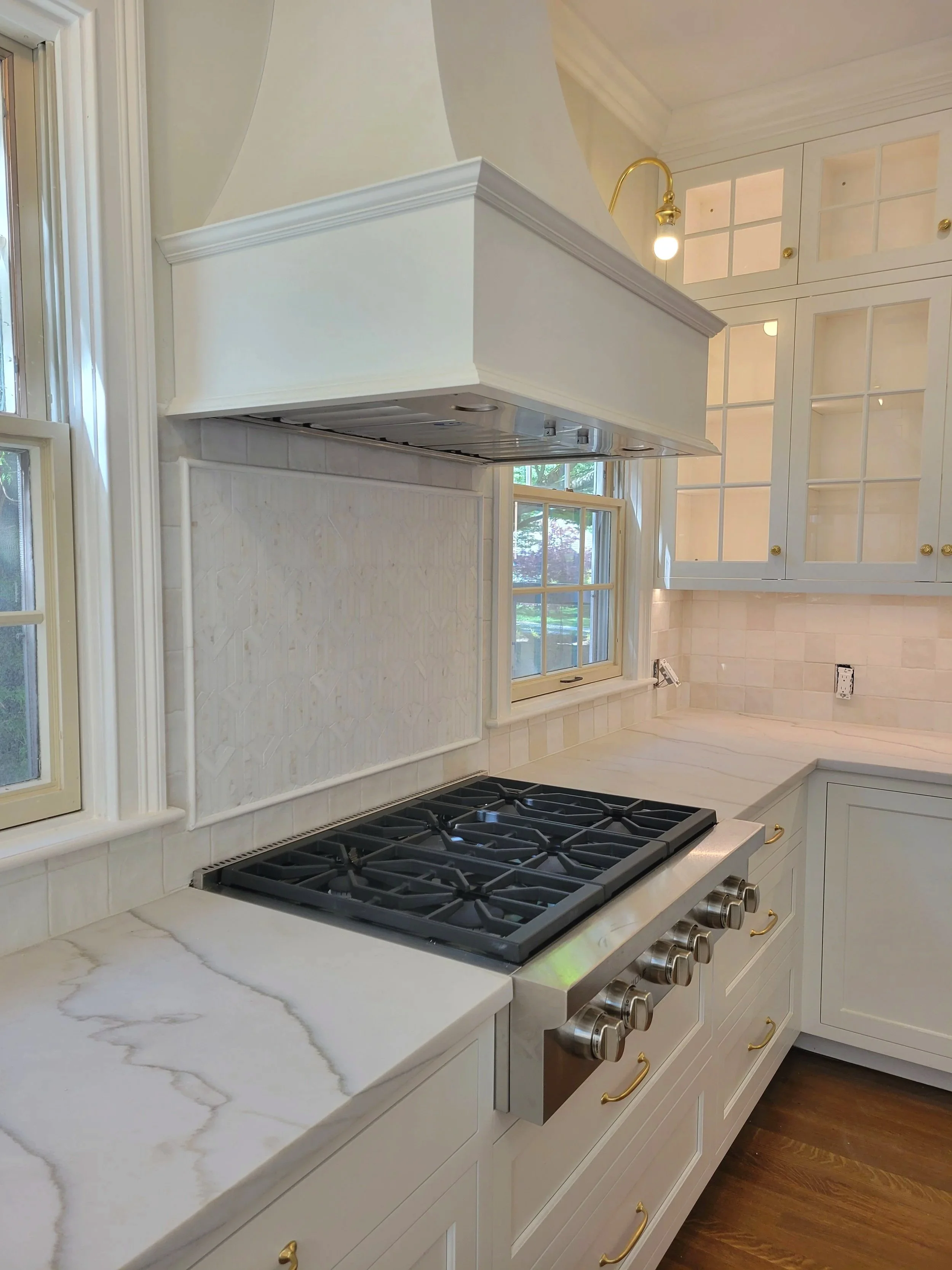 A kitchen with a white marble countertop, gas stove with four burners, white cabinets with glass doors and gold knobs, a window with a view outside, and a range hood above the stove.