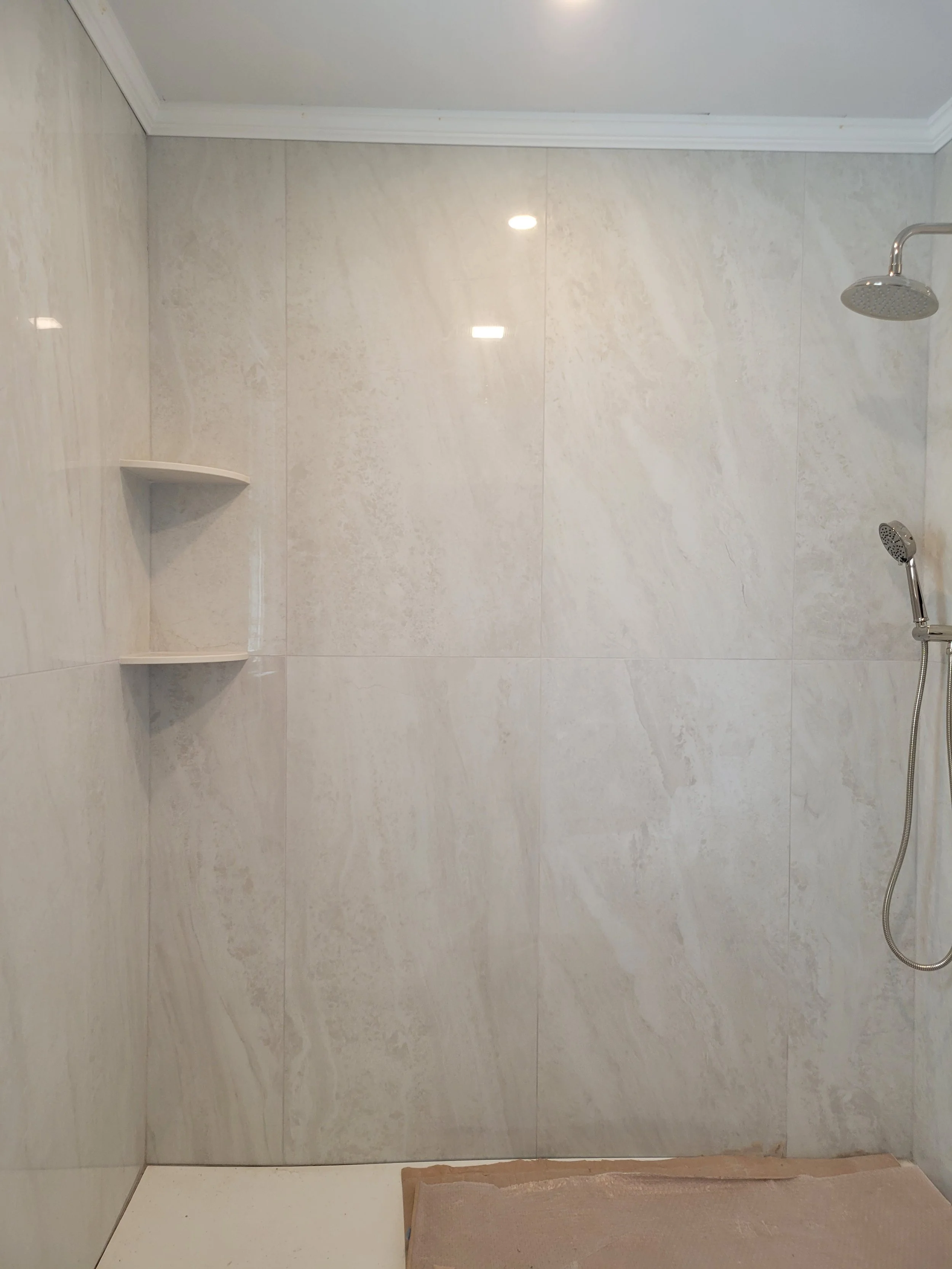 A walk-in shower with beige marble tiles, a showerhead on the right, and a built-in corner shelf on the left wall.