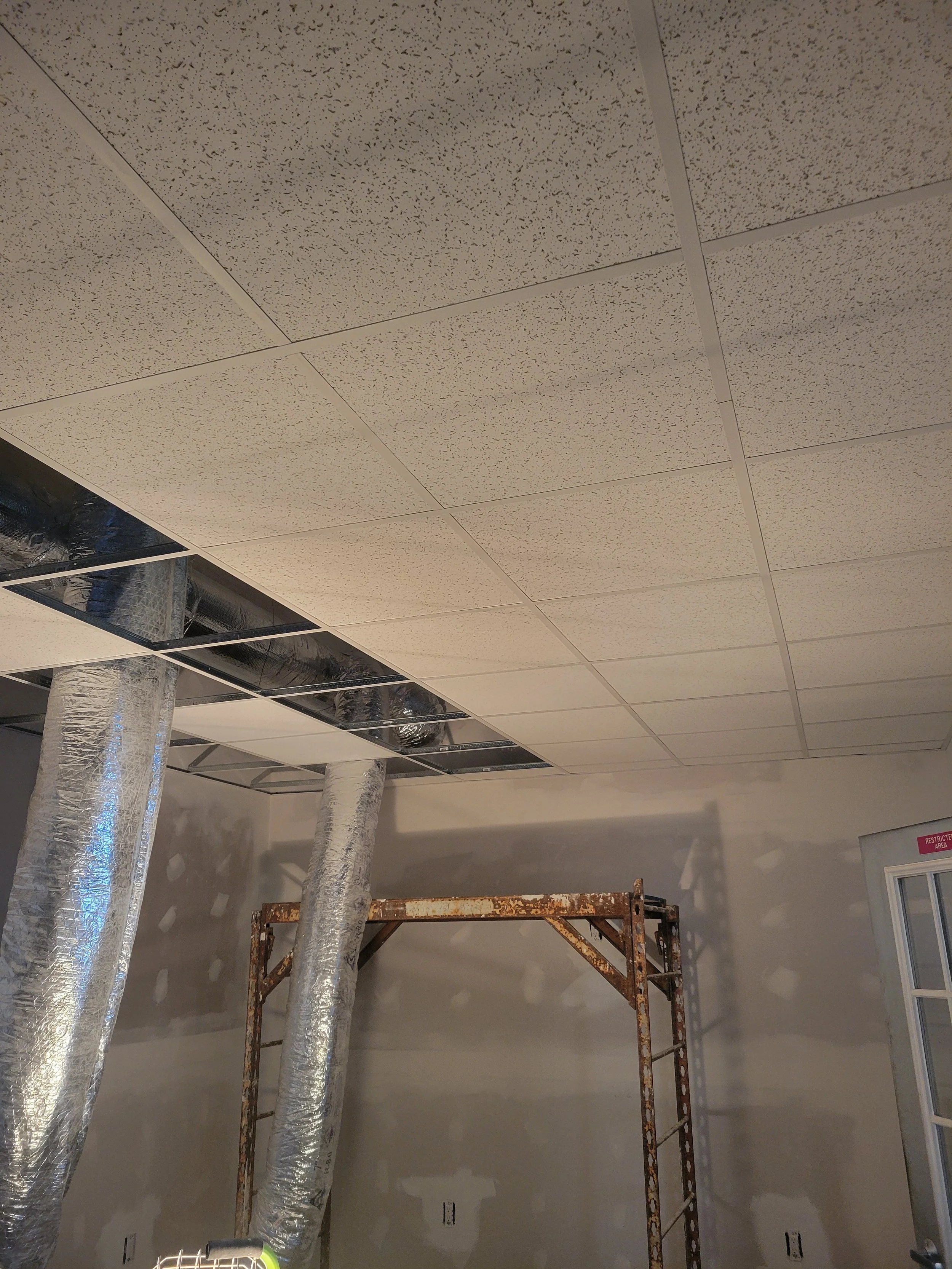 Ceiling tiles with exposed ductwork, insulation pipes, and construction scaffolding in an unfinished room.