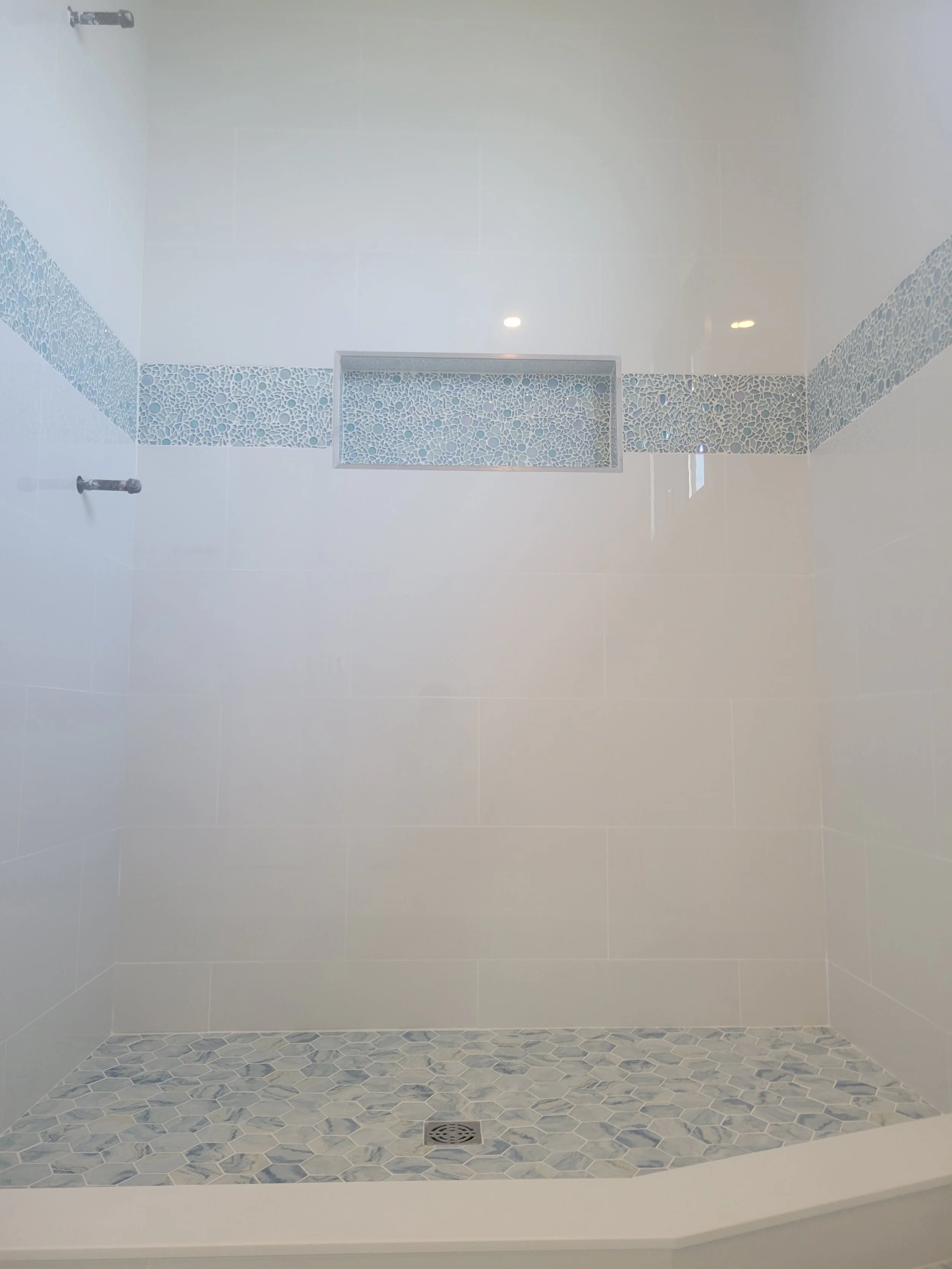 A modern walk-in shower with white walls, a decorative mosaic tile band, a linear drain, and hexagonal floor tiles in various shades of blue and gray.