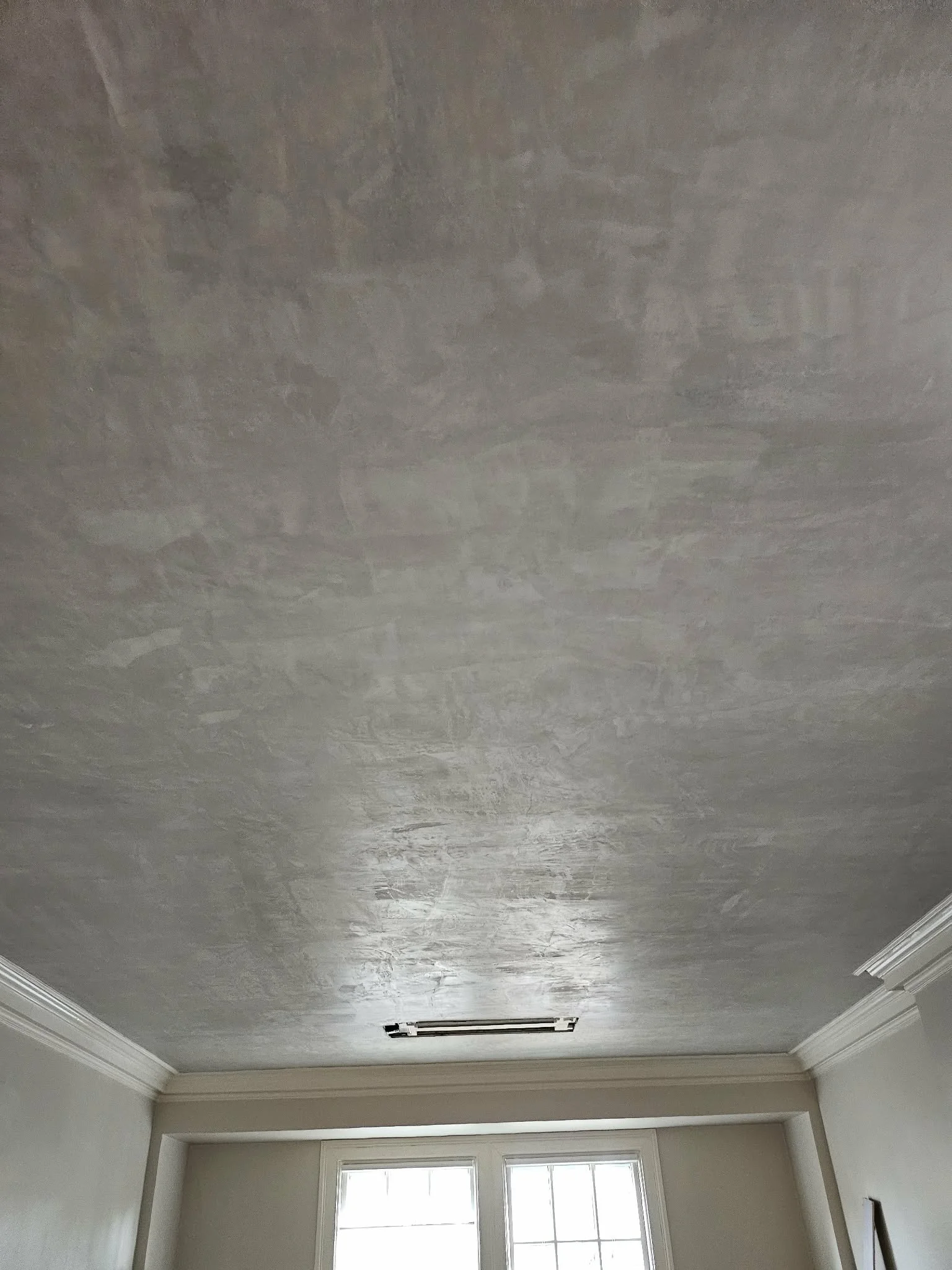 The ceiling of a room with an unfinished or textured gray surface, a white crown molding, a ceiling vent, and a window letting in natural light.