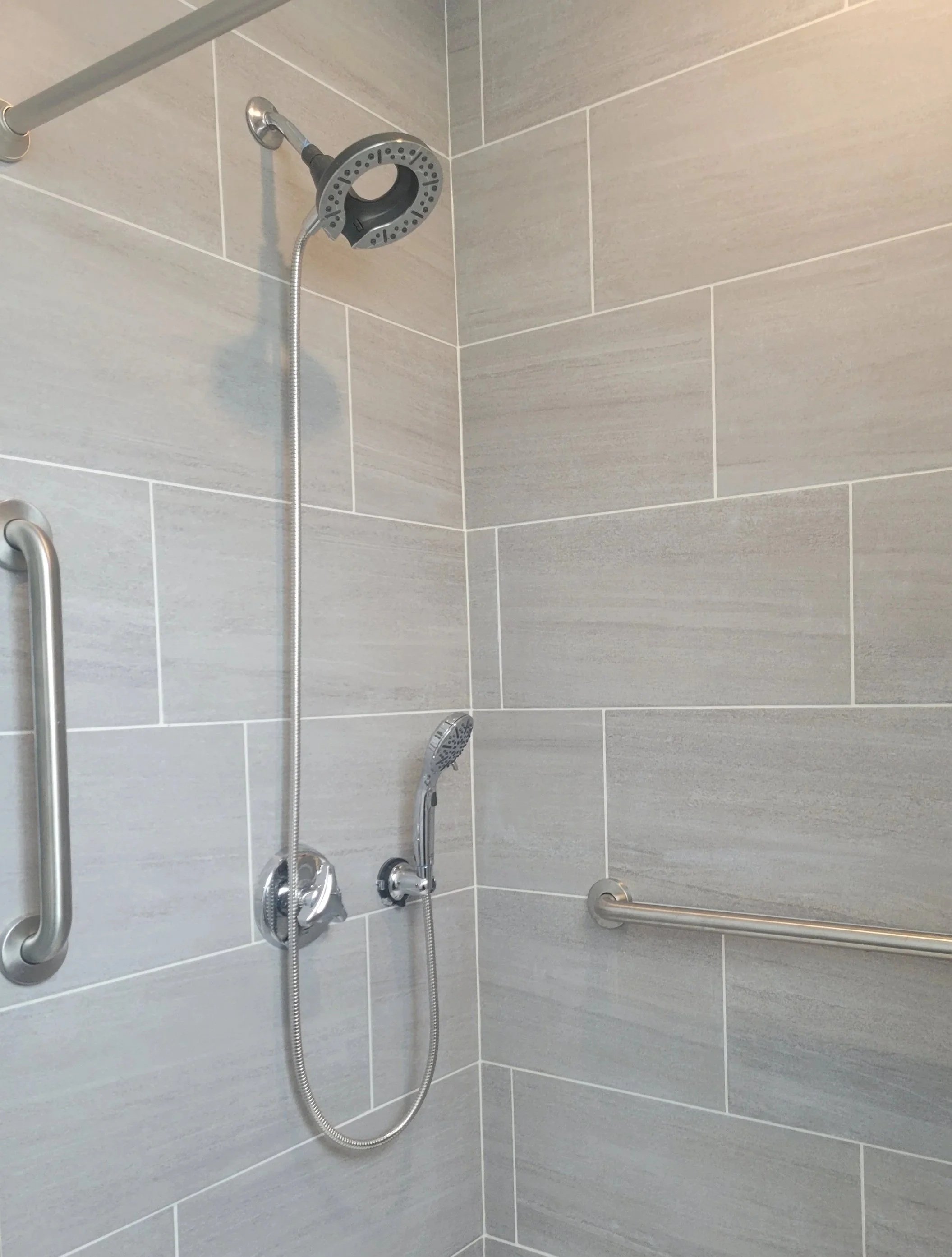 An accessible shower with beige tiled walls, a handheld showerhead, and safety grab bars.