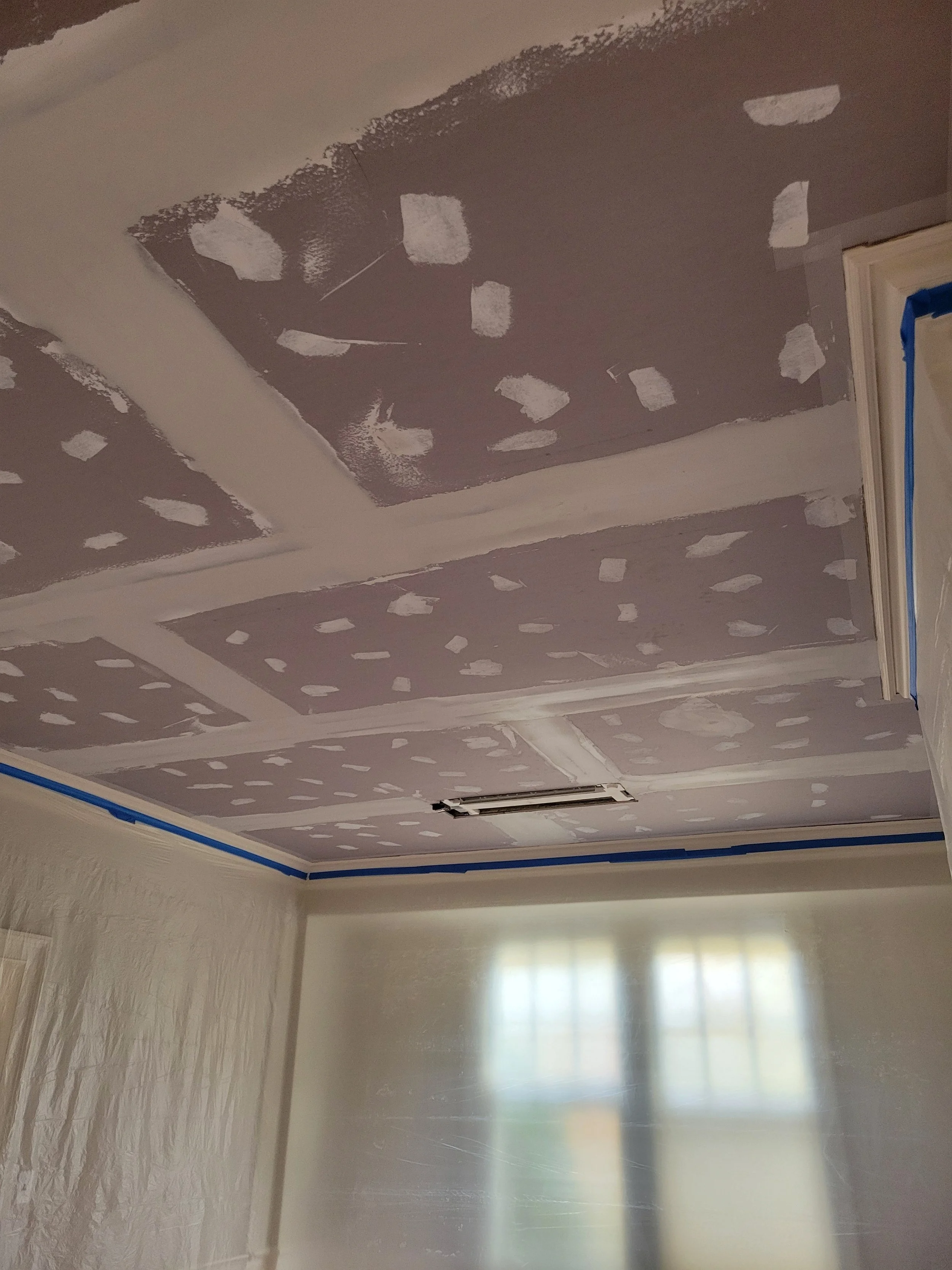 Ceiling with drywall patches and taped seams in the process of being painted or finished, with a window below and protective plastic on the walls.