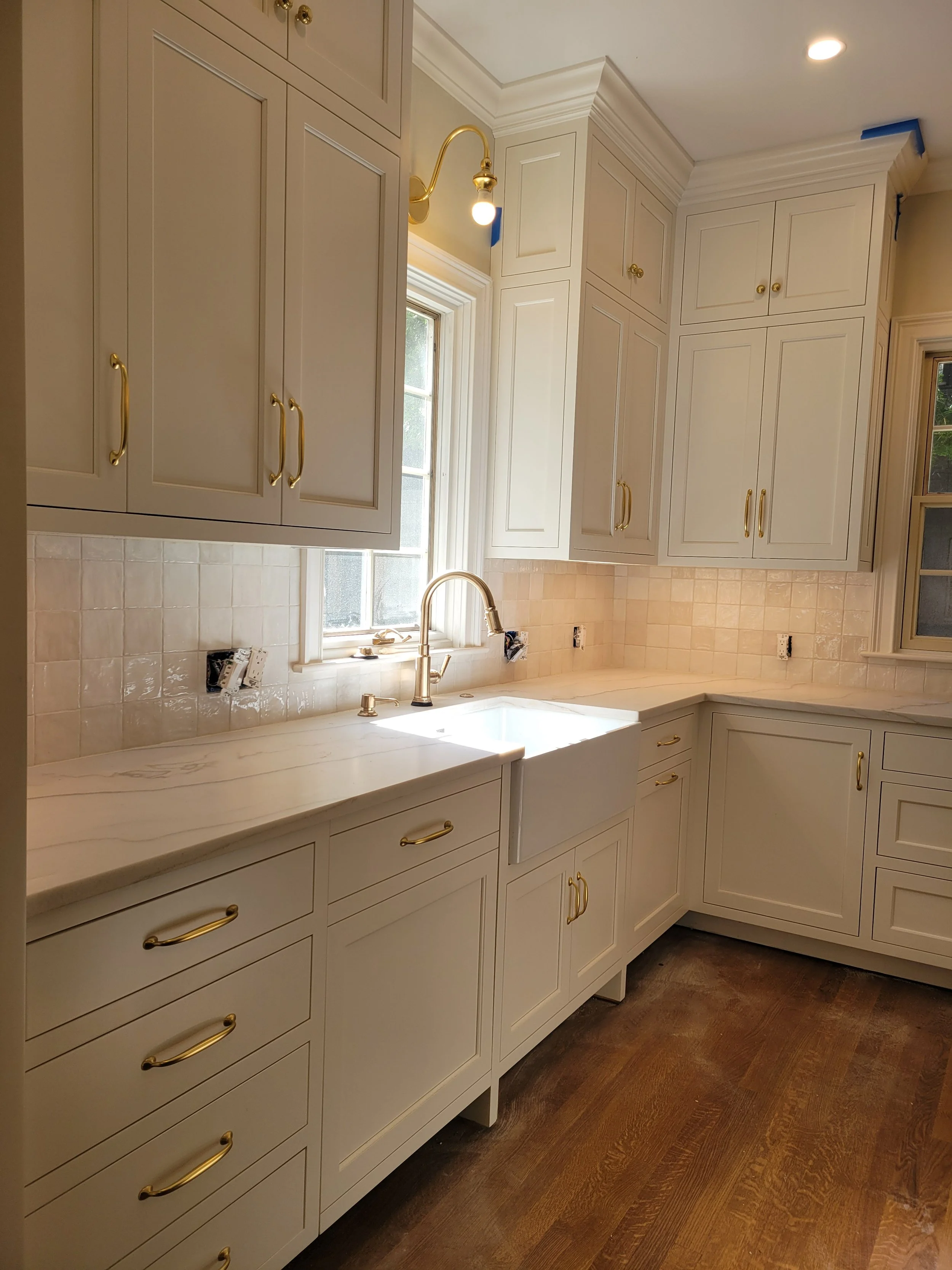 Kitchen with white cabinets, gold handles, beige tiled backsplash, and a window above the sink with a gold faucet. Some electrical outlets are visible on the backsplash, and the floor is wooden.
