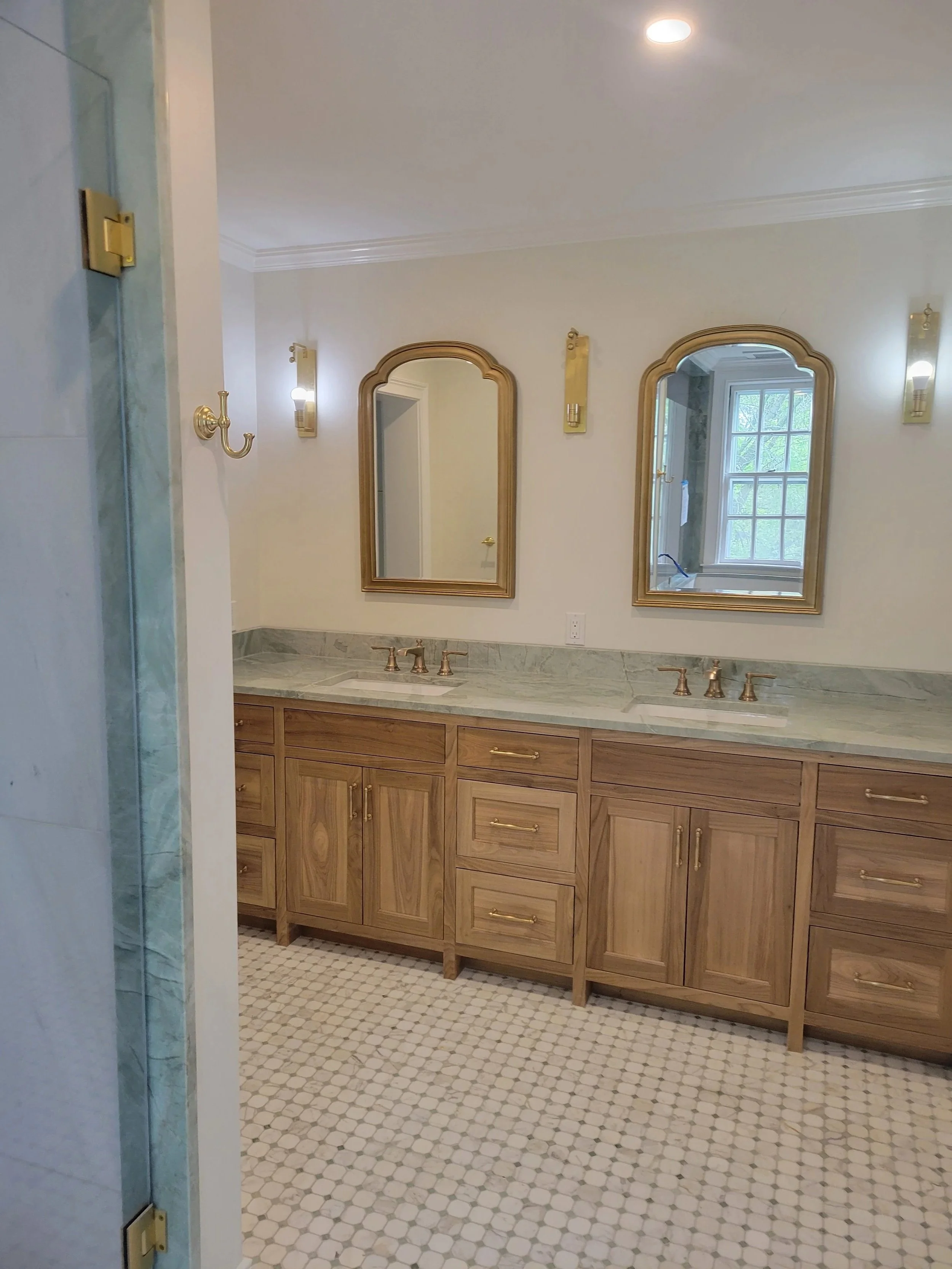 A bathroom interior with a wooden vanity, two mirrors with arched wooden frames, and gold fixtures. The floor features small hexagon tiles, and there are wall-mounted lights and a window with a view of greenery outside.