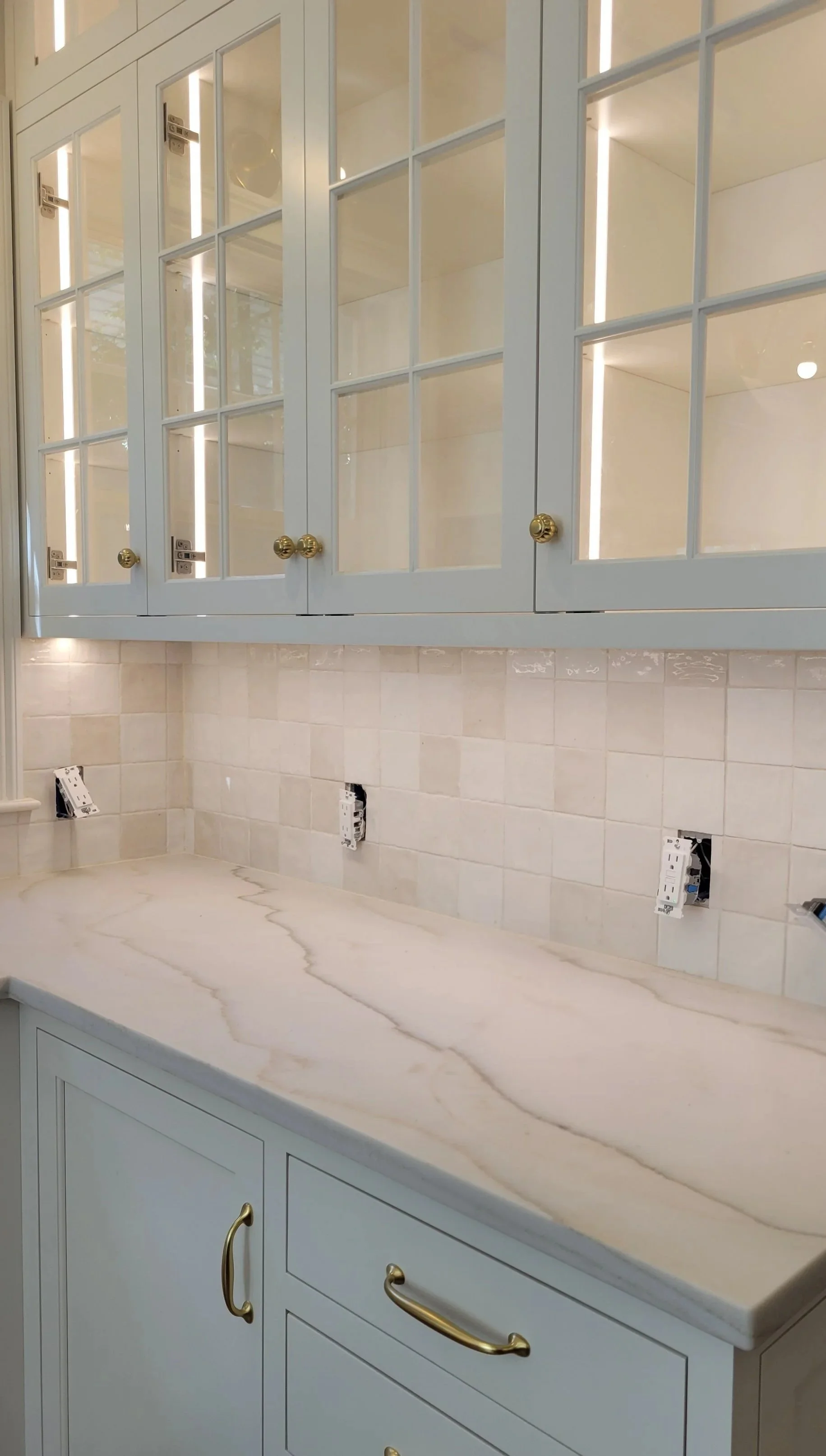Kitchen with light blue cabinets with glass-paneled doors, gold knobs, a beige marble countertop with gray veining, and beige tiled backsplash with electrical outlets.