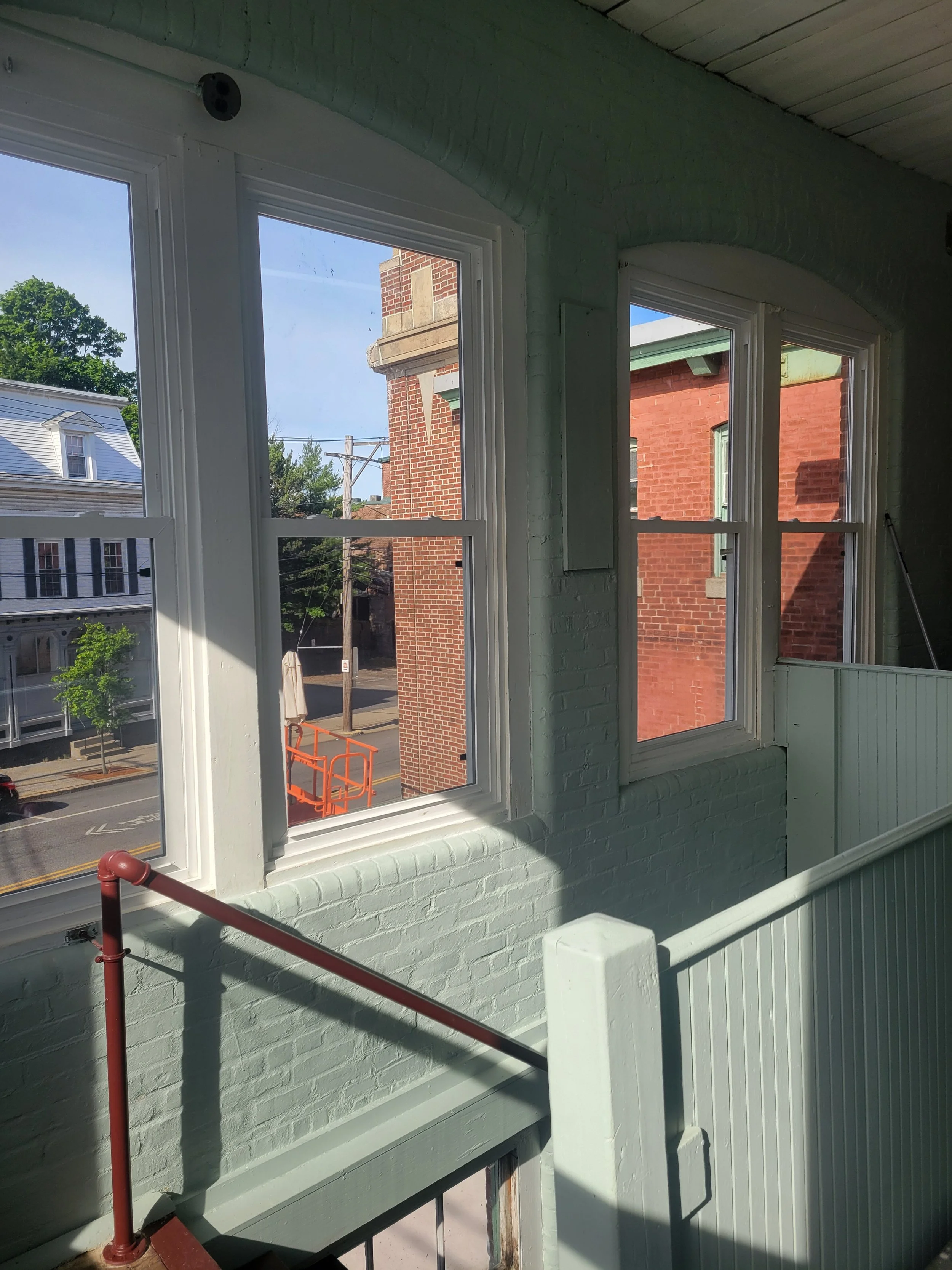 Interior view of a building with three large windows showing an exterior street scene with a brick building, trees, and a metal basketball hoop.