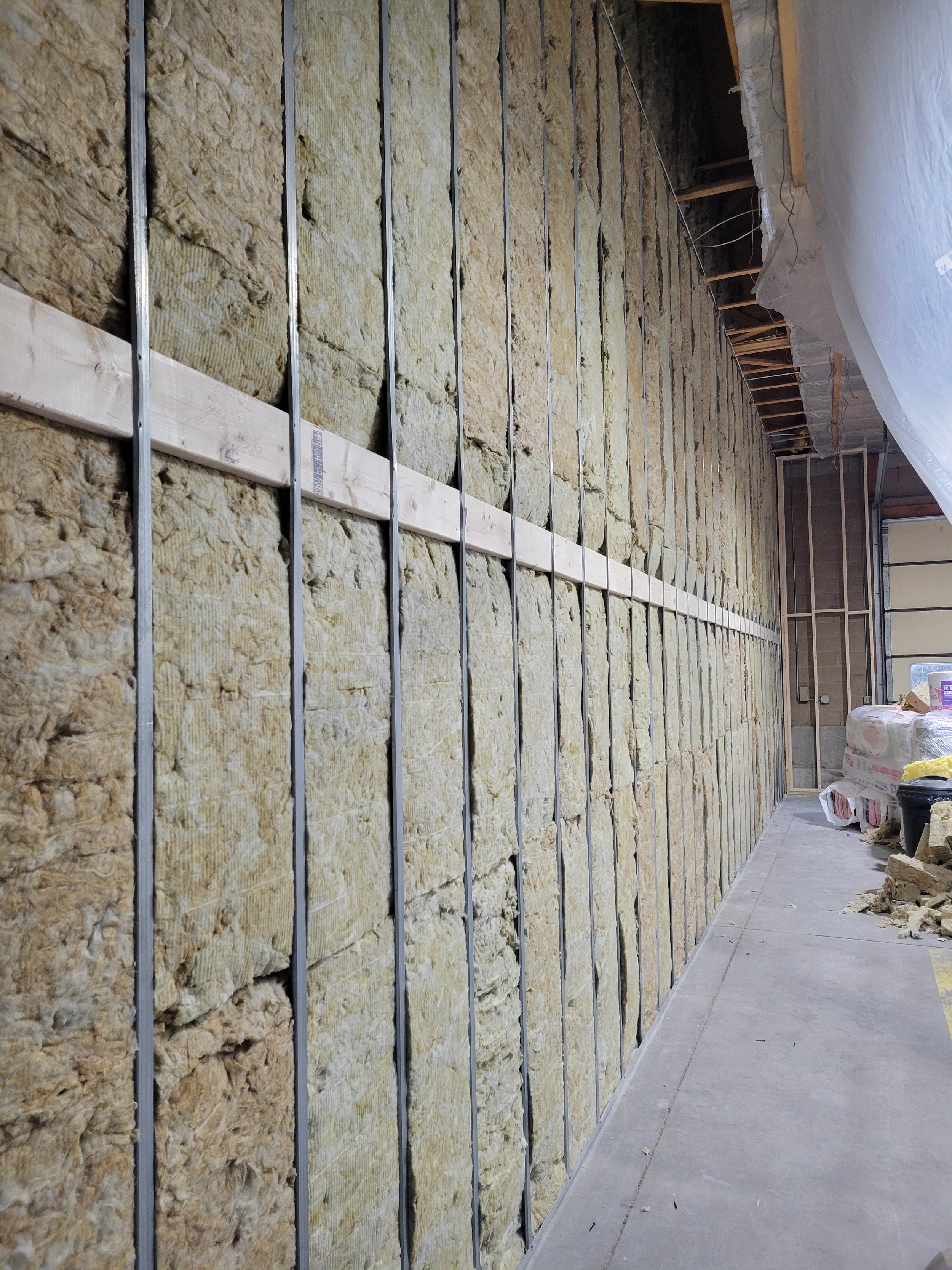 Interior of a building under construction with a wall being insulated with spray foam insulation. Metal framing studs are visible, and construction materials and tools are on the floor.