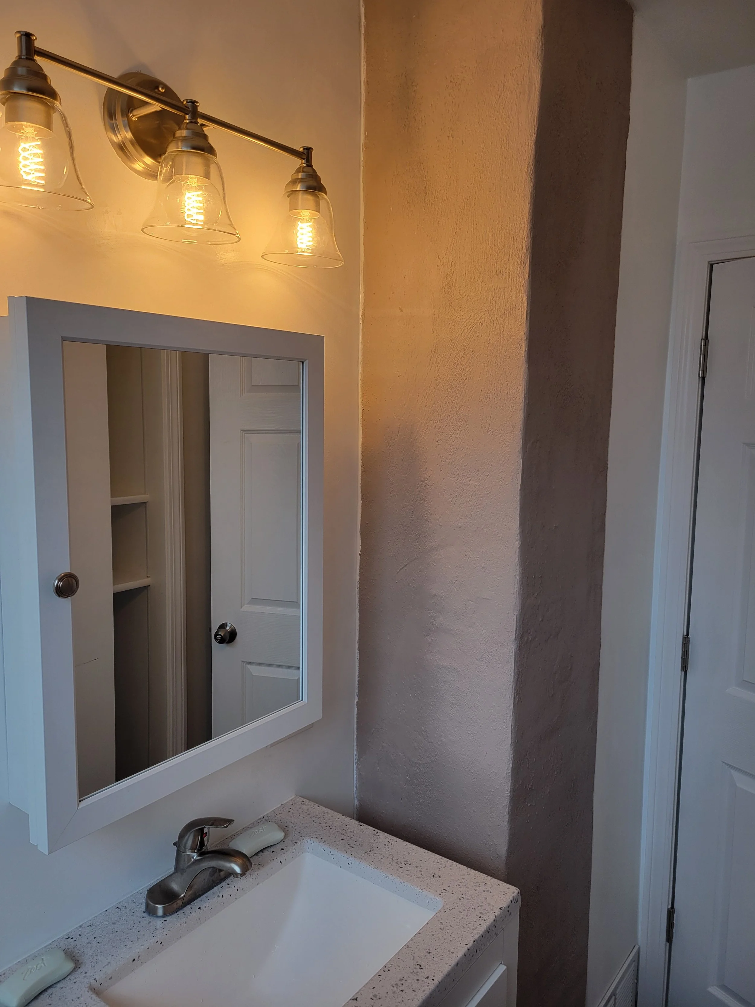 Bathroom vanity with white countertop, undermount sink, and a brushed nickel faucet. Above the sink is a wall-mounted cabinet with a mirror, and a three-light fixture with glass shades is mounted on the wall above the mirror. To the right, there is a textured wall and a door with visible hinges.
