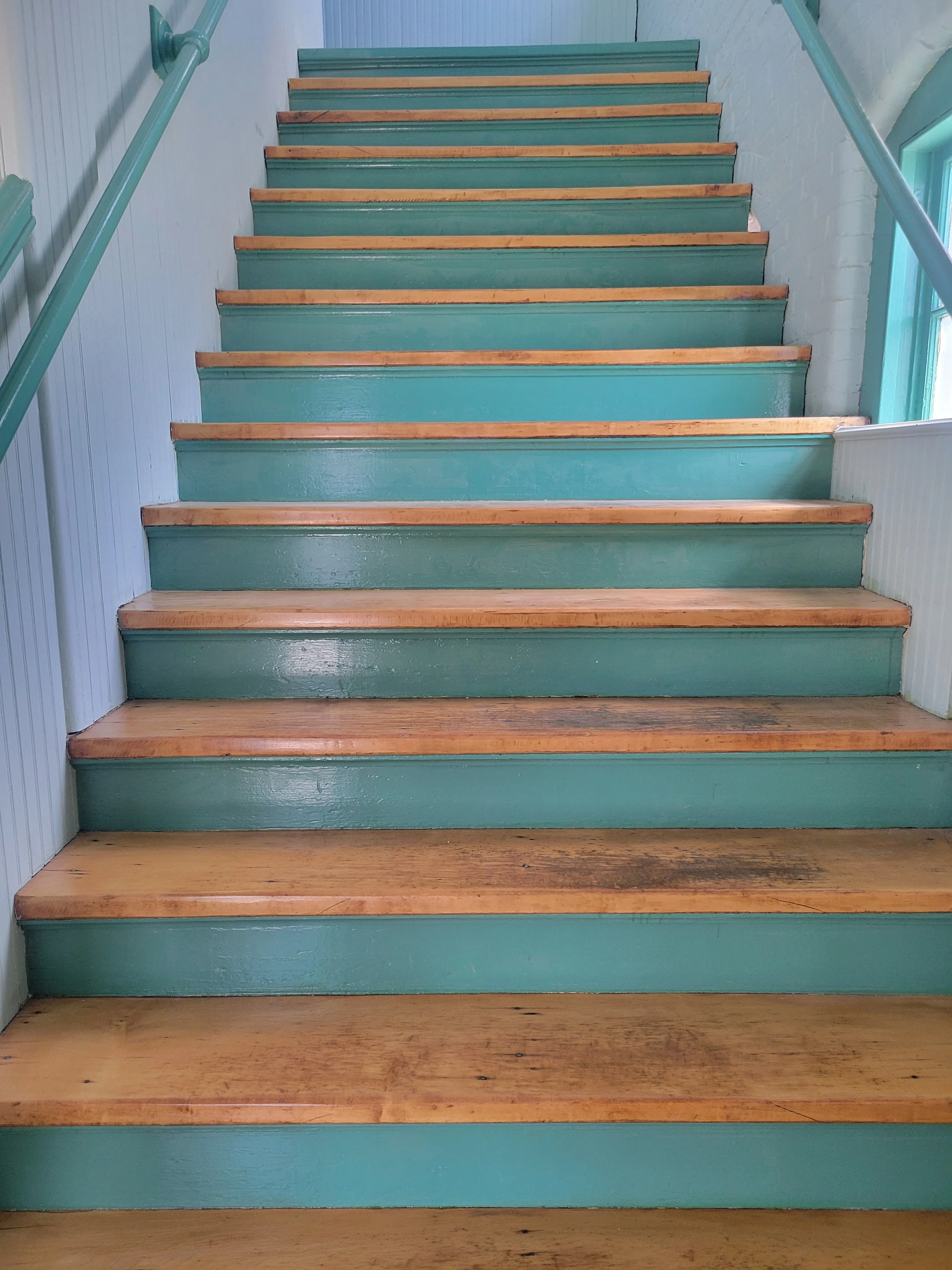 Wooden stairs with teal risers and natural wood treads, flanked by teal handrails and wall paneling, leading upward near a window.