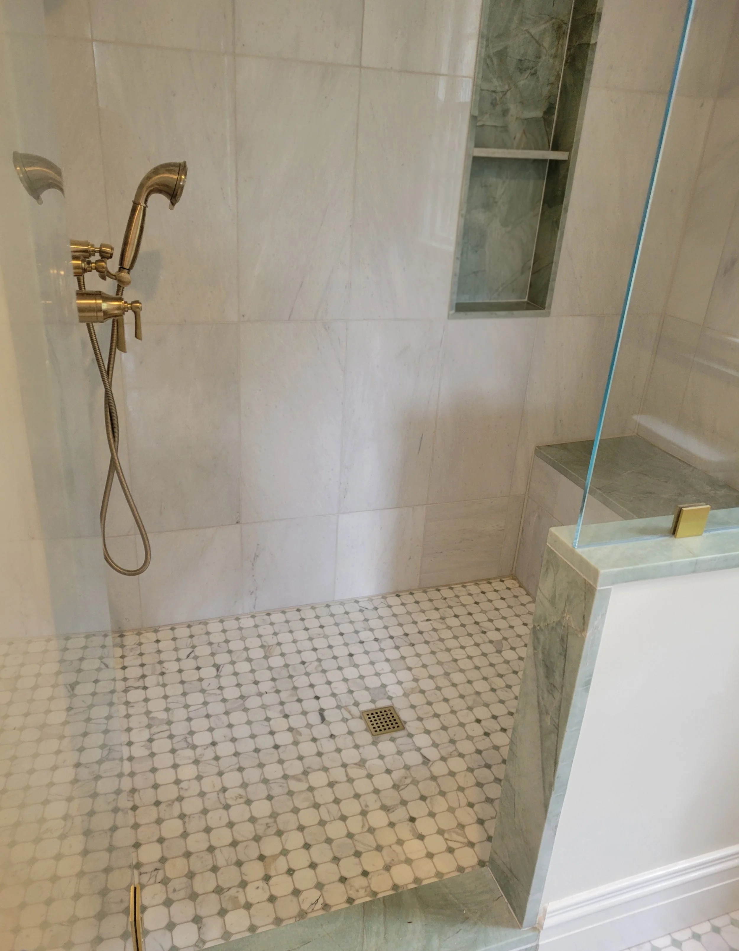 Bathroom shower area with beige marble walls, a built-in shower niche, a brass shower head and fixtures, and a white tile floor with small hexagonal tiles.