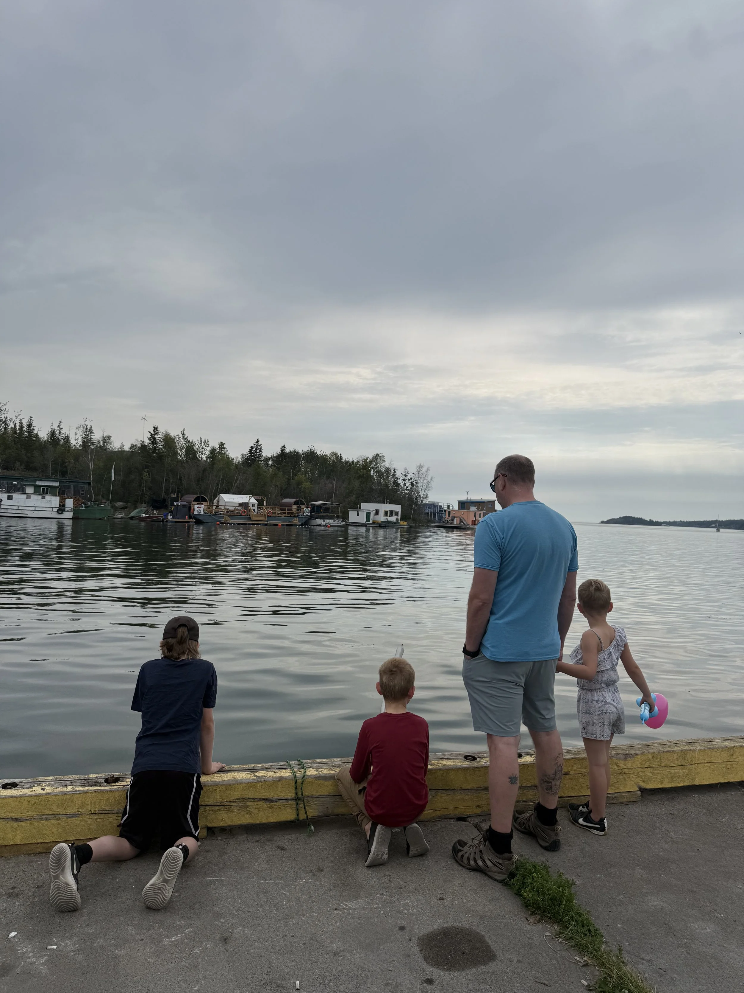People standing and sitting near the water, observing boats and houseboats on a cloudy day