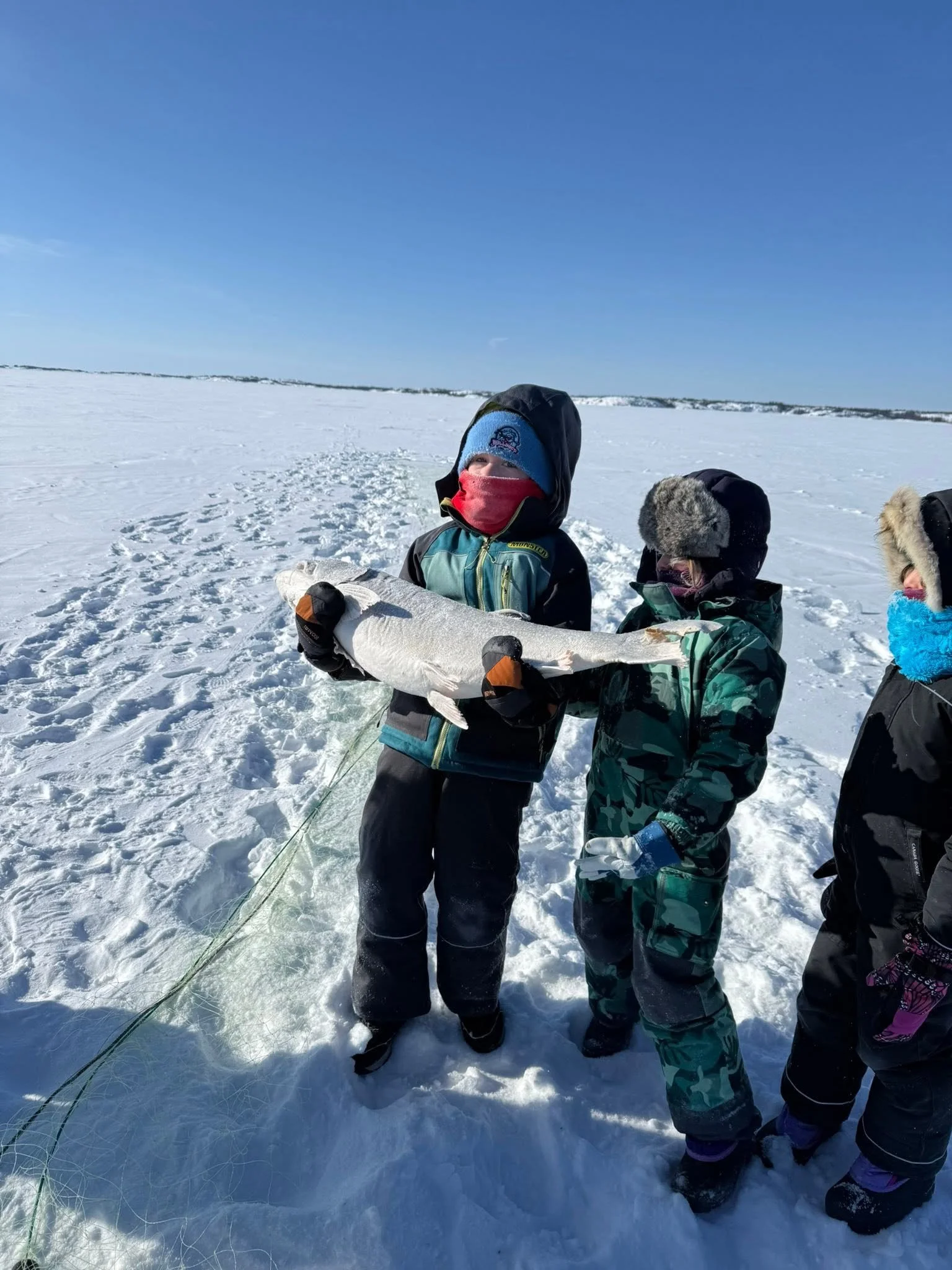 Children dressed in winter clothing standing on snow, holding a large fish they caught, with a snowy landscape and a clear blue sky in the background.