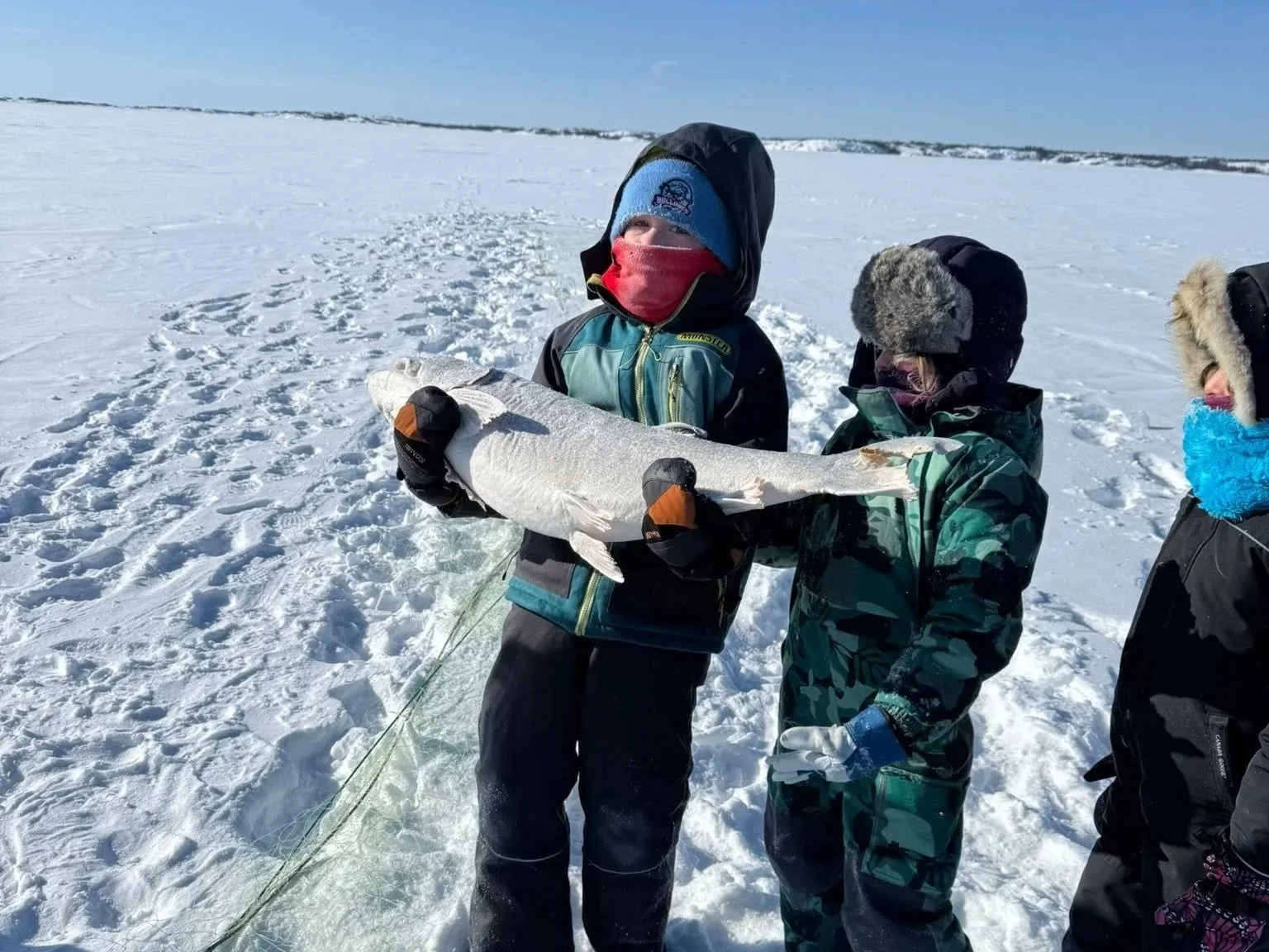 Three children dressed in winter clothing standing on snow-covered ice or snow field, holding a large fish, with a wide open snow landscape and cloudy sky in the background.