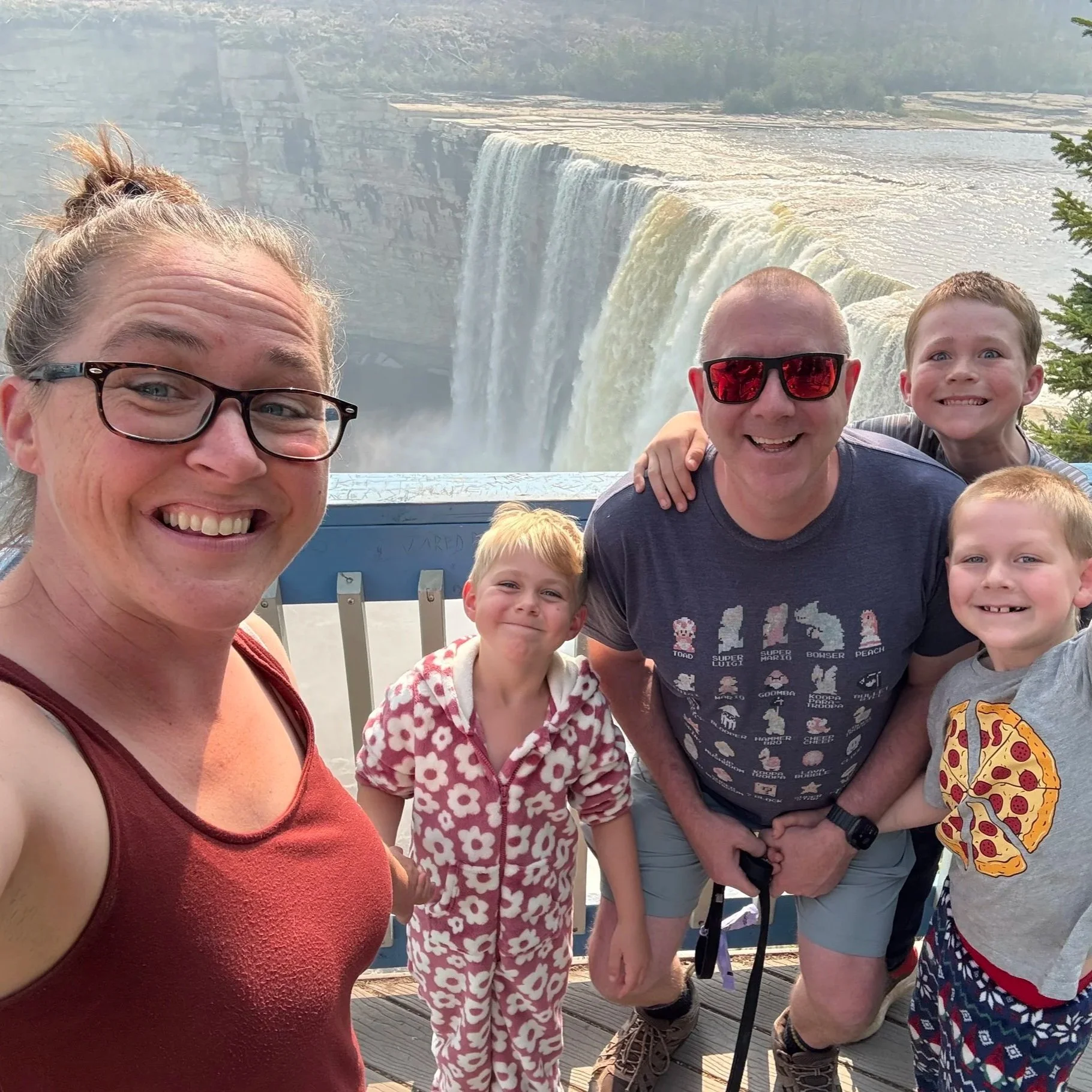 Family of five taking a selfie at Niagara Falls with the waterfall in the background. The group includes three children and two adults, all smiling and wearing casual clothing, with one adult wearing sunglasses and the other a tank top.