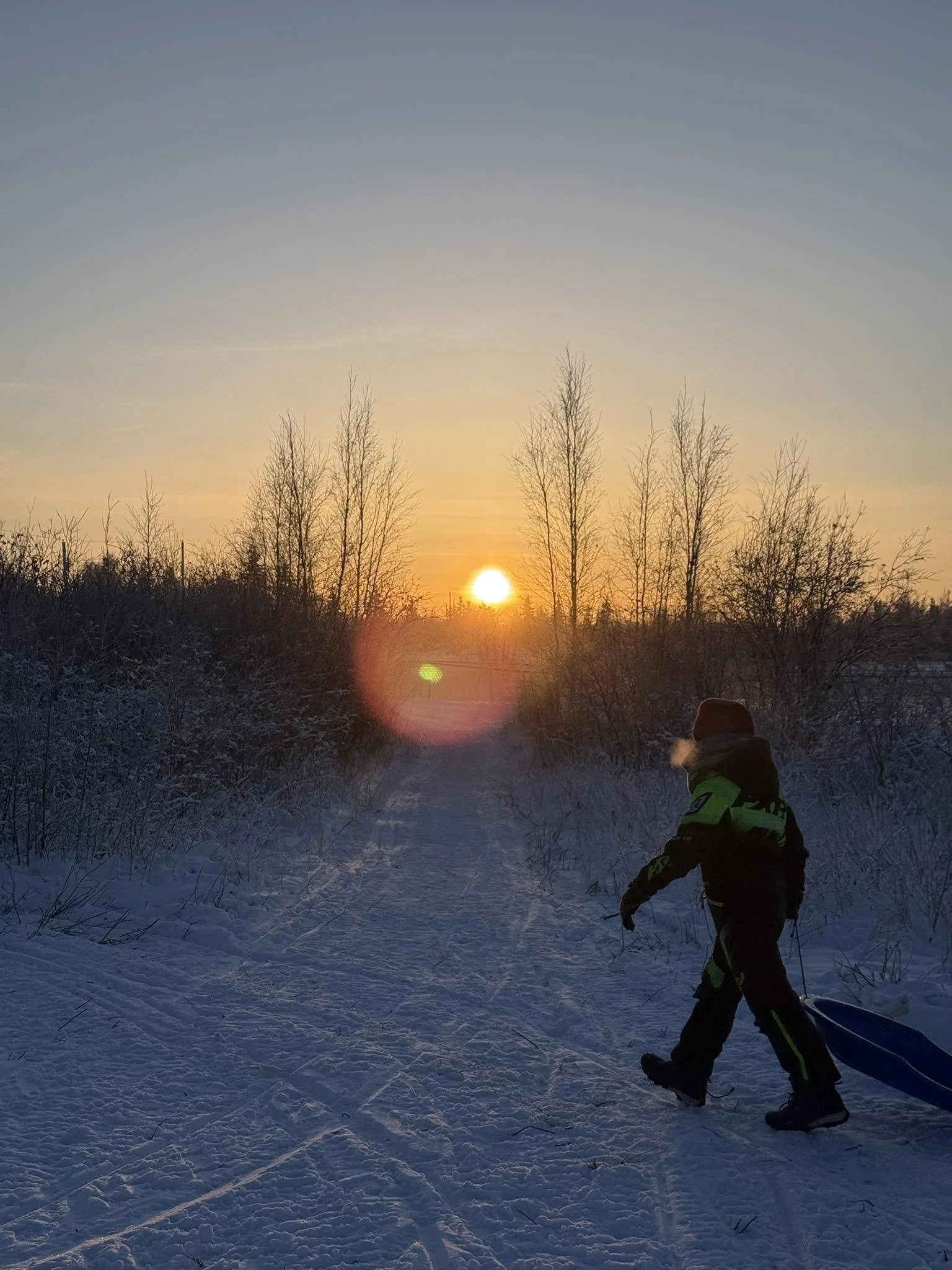A child dressed in winter clothing, including a helmet and gloves, is walking on a snowy path during sunrise or sunset. The sky is clear with a soft gradient of colors, and bare trees line the horizon.