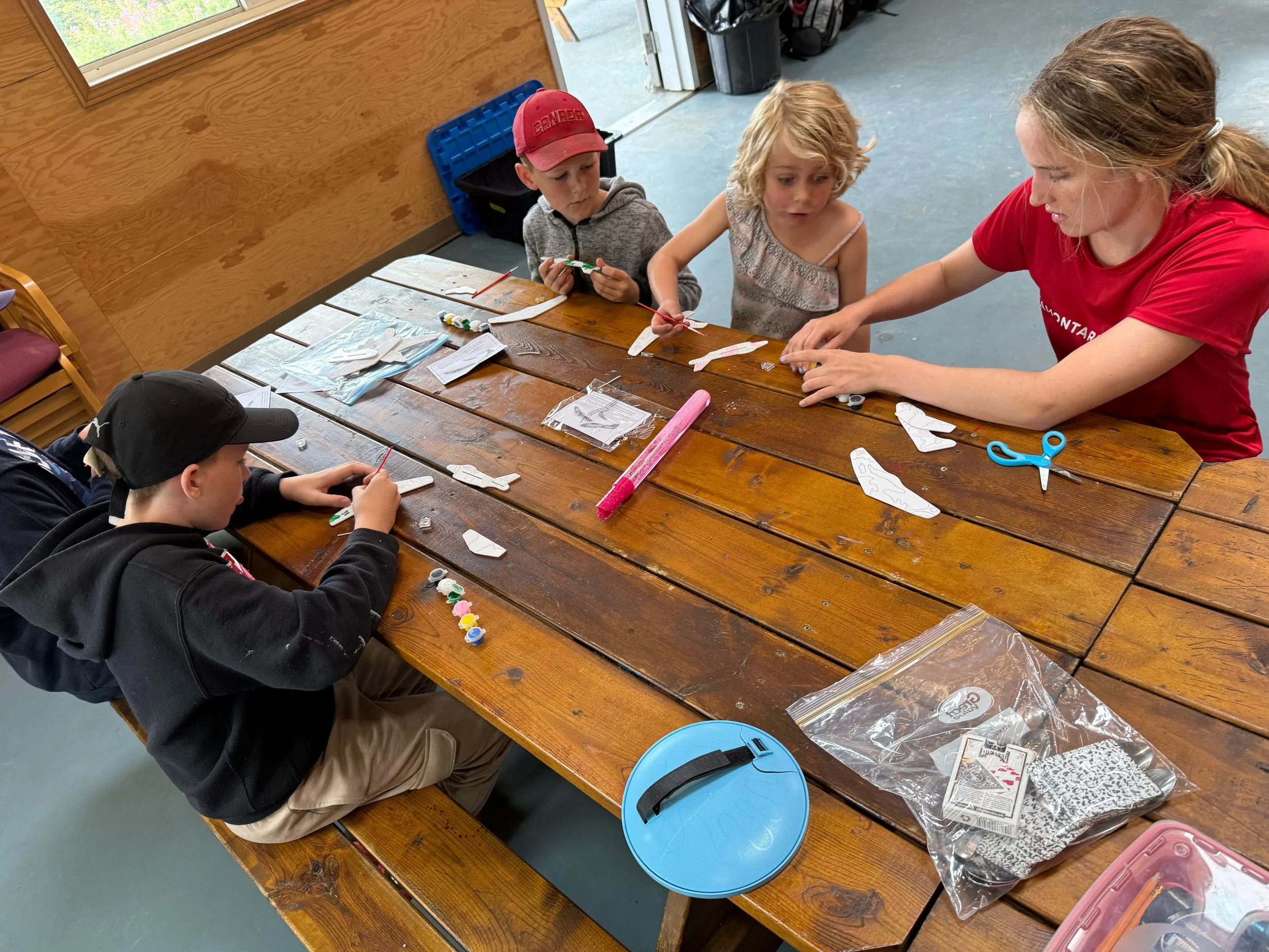 Children and an adult making paper airplanes with craft supplies on a wooden table inside a room.