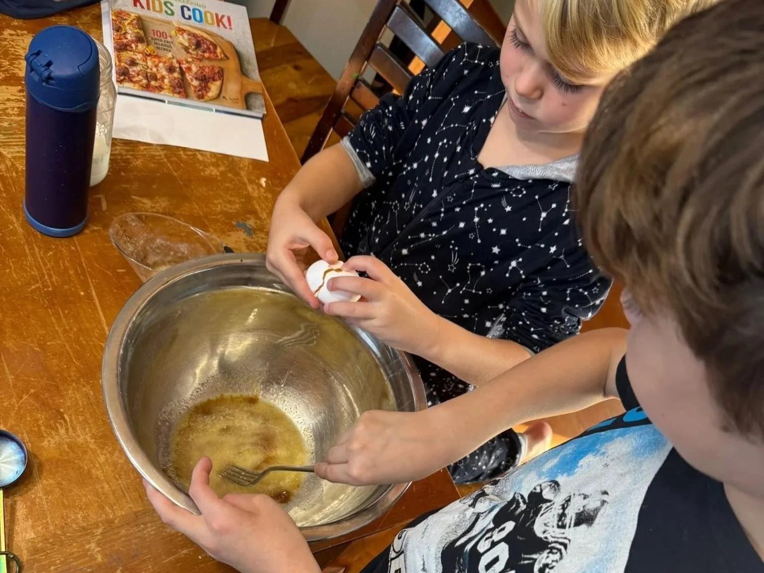 Two children cracking eggs into a mixing bowl on a wooden table, with a pizza magazine and water bottle nearby.