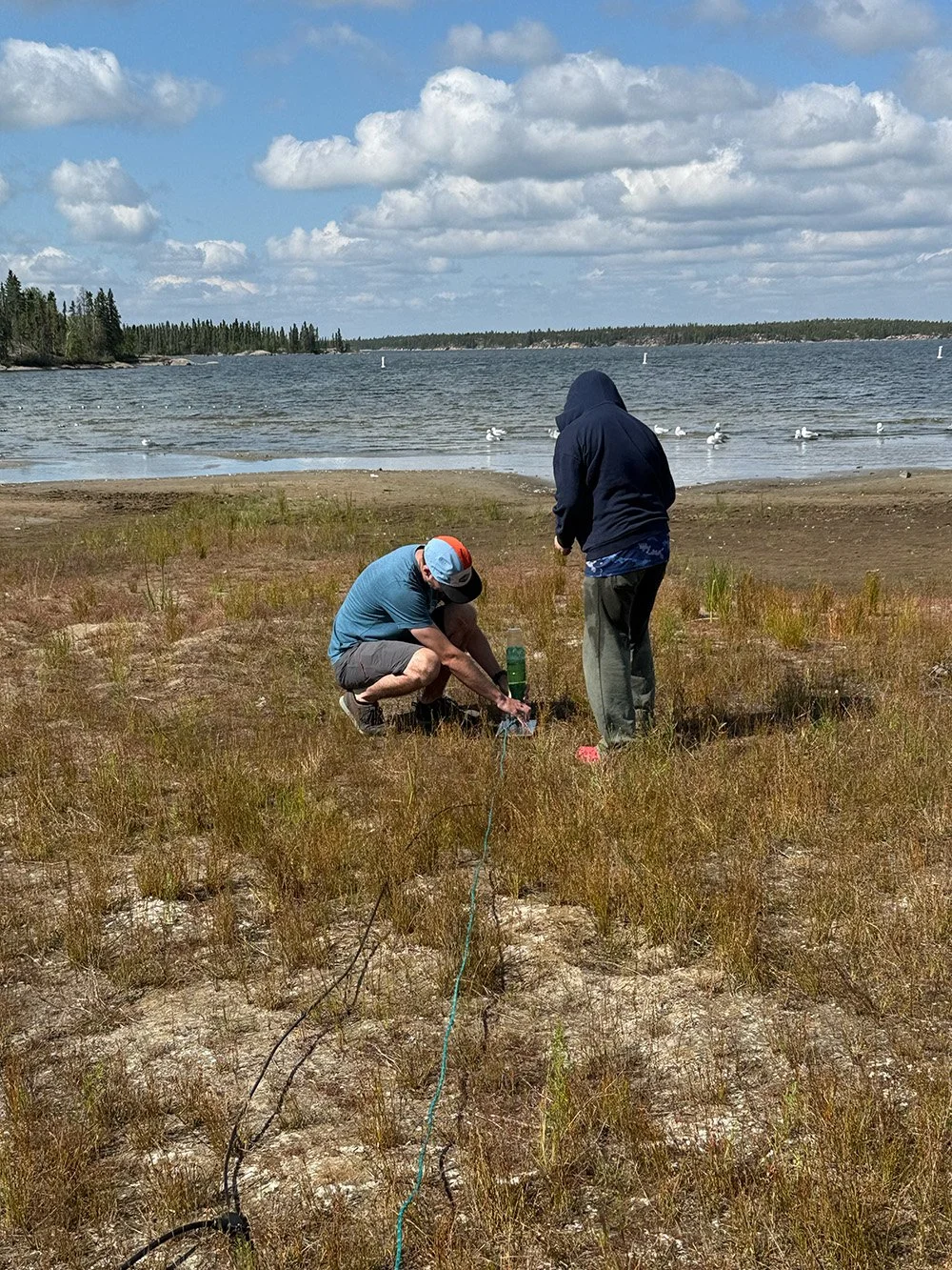 Two men on a sandy area near a body of water, one is crouched down and working with equipment, the other is standing nearby, with seagulls flying over the water in the background and a partly cloudy sky above.
