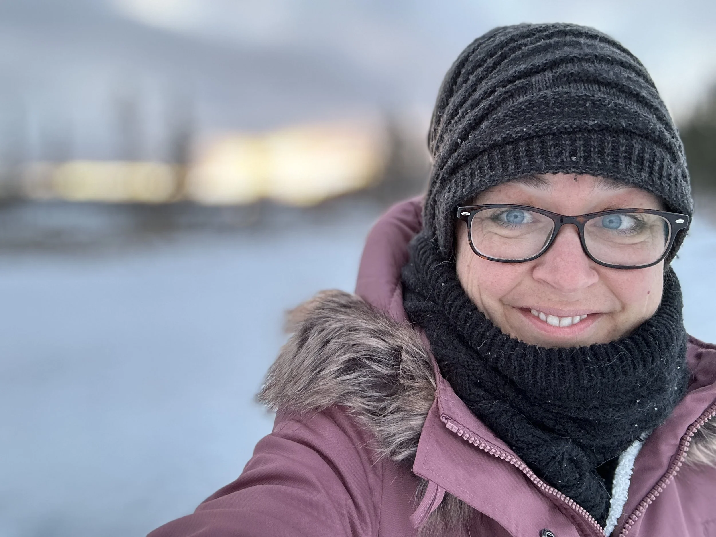 Close-up of a smiling woman wearing a gray knit beanie, glasses, and a pink winter coat with a fur-lined hood. She is outdoors on a cloudy day.