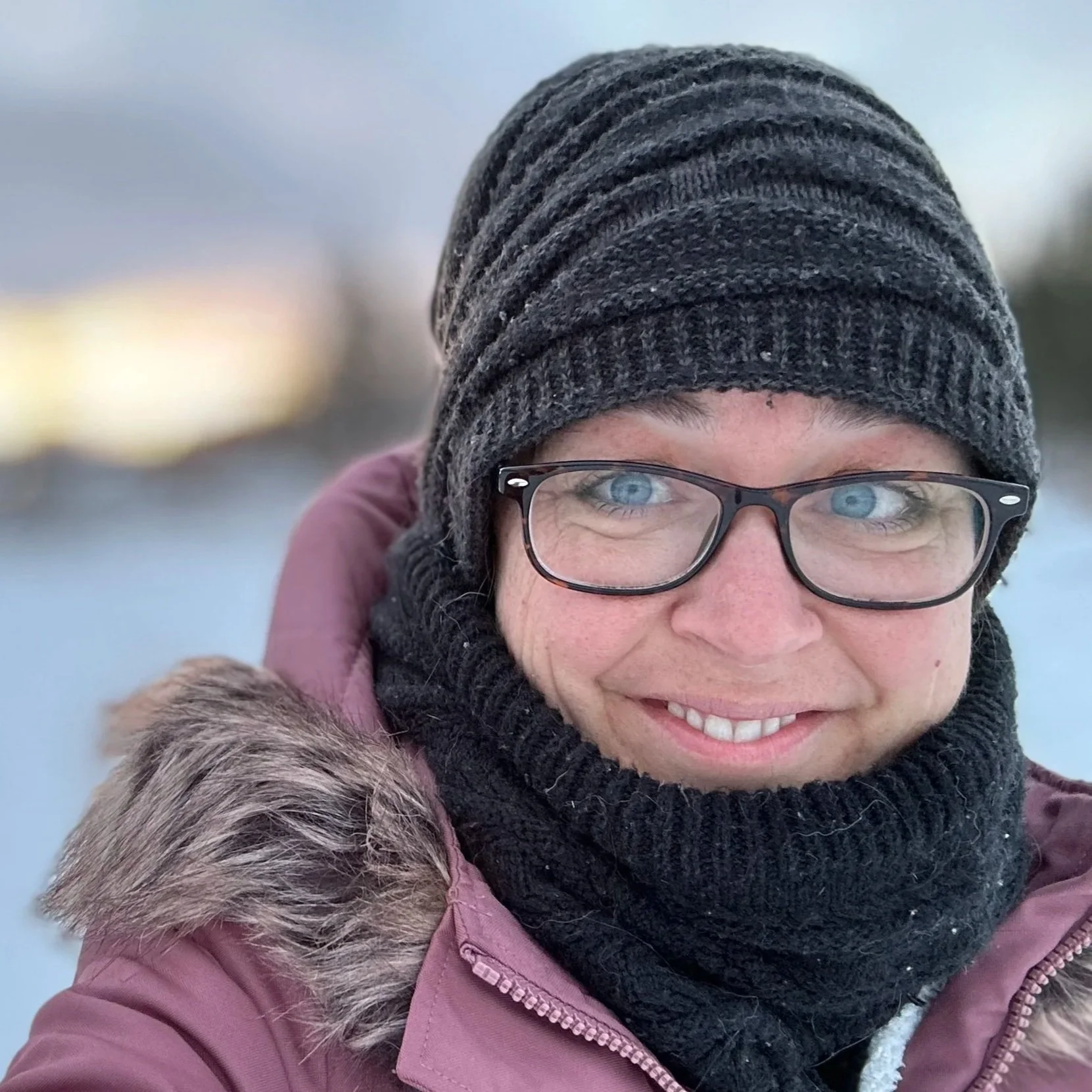 A smiling person outdoors in winter, wearing a gray knit beanie, black glasses, a black scarf, and a pink coat with fur trim.