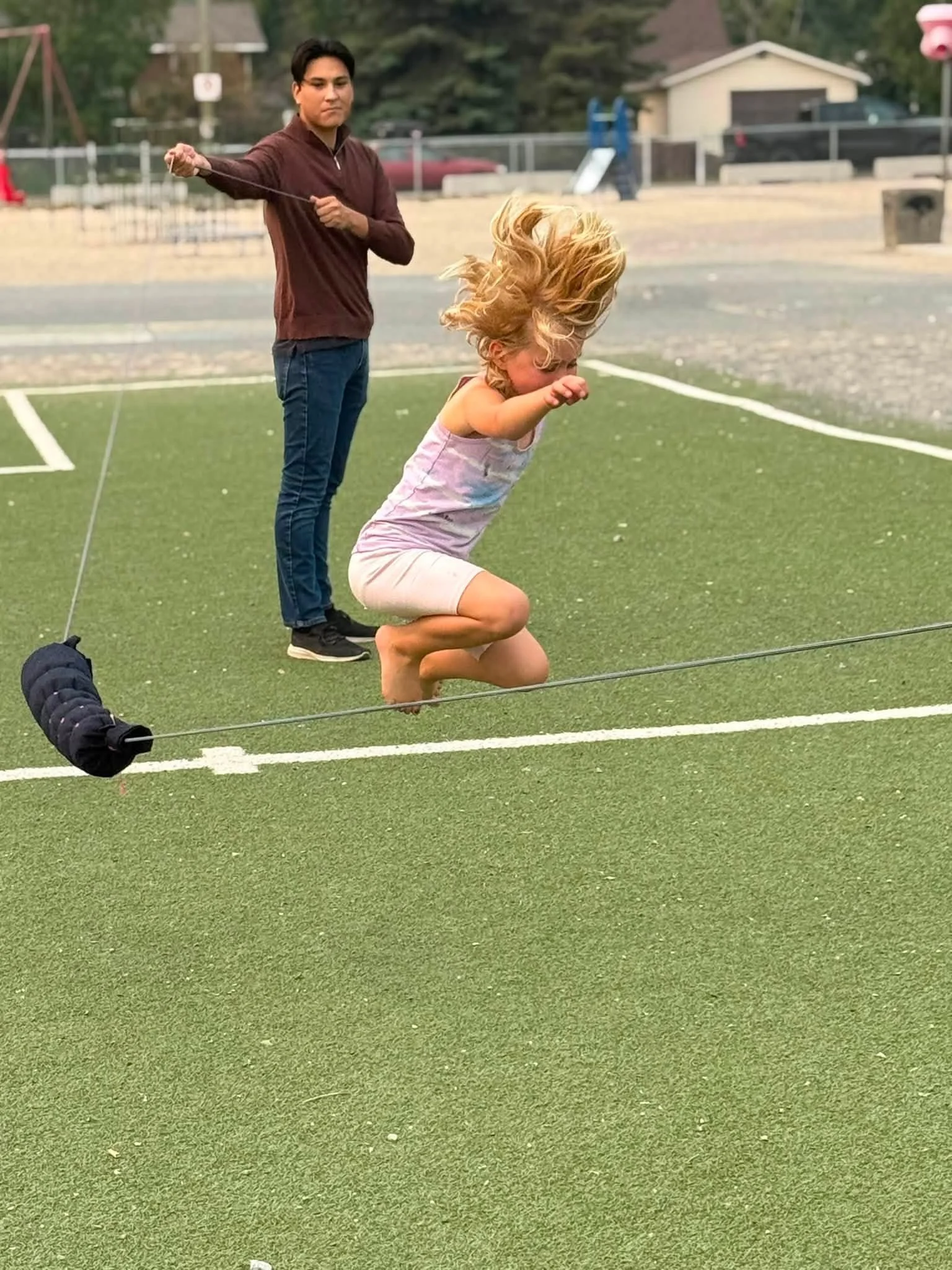 A woman kneels on a green field while a man stands behind her holding a string connected to a slackline, with her hair blowing in the wind.