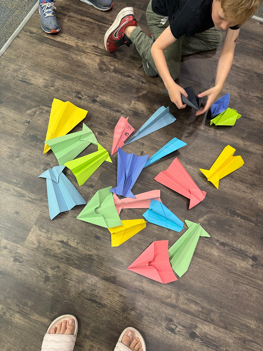 A group of colorful paper paper airplanes arranged on a wooden floor, with a child in black shirt and green pants playing with one paper airplane.