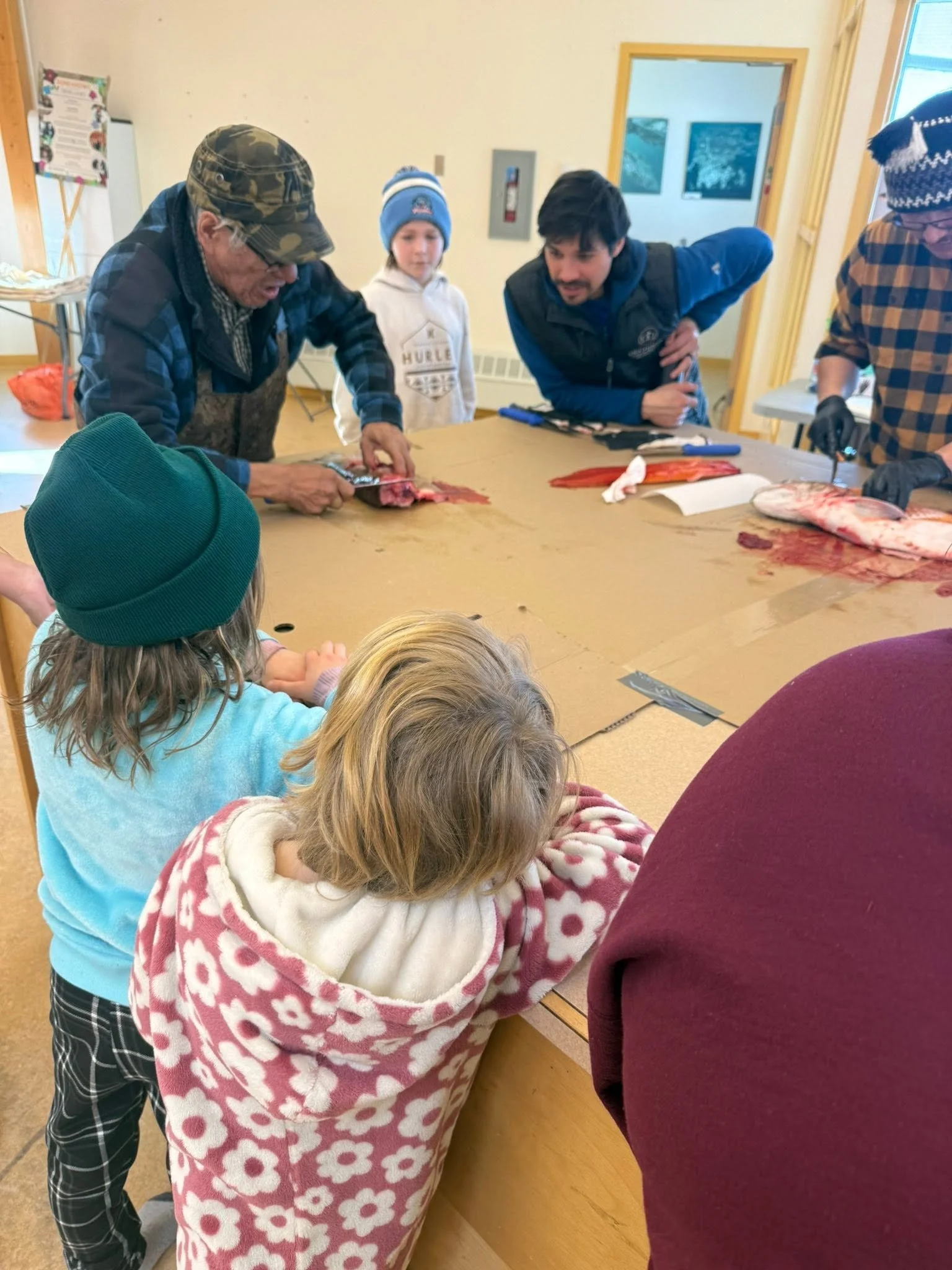 People gathered around a table, with one man demonstrating fish cleaning, a young girl observing, and others watching, in an indoor setting.