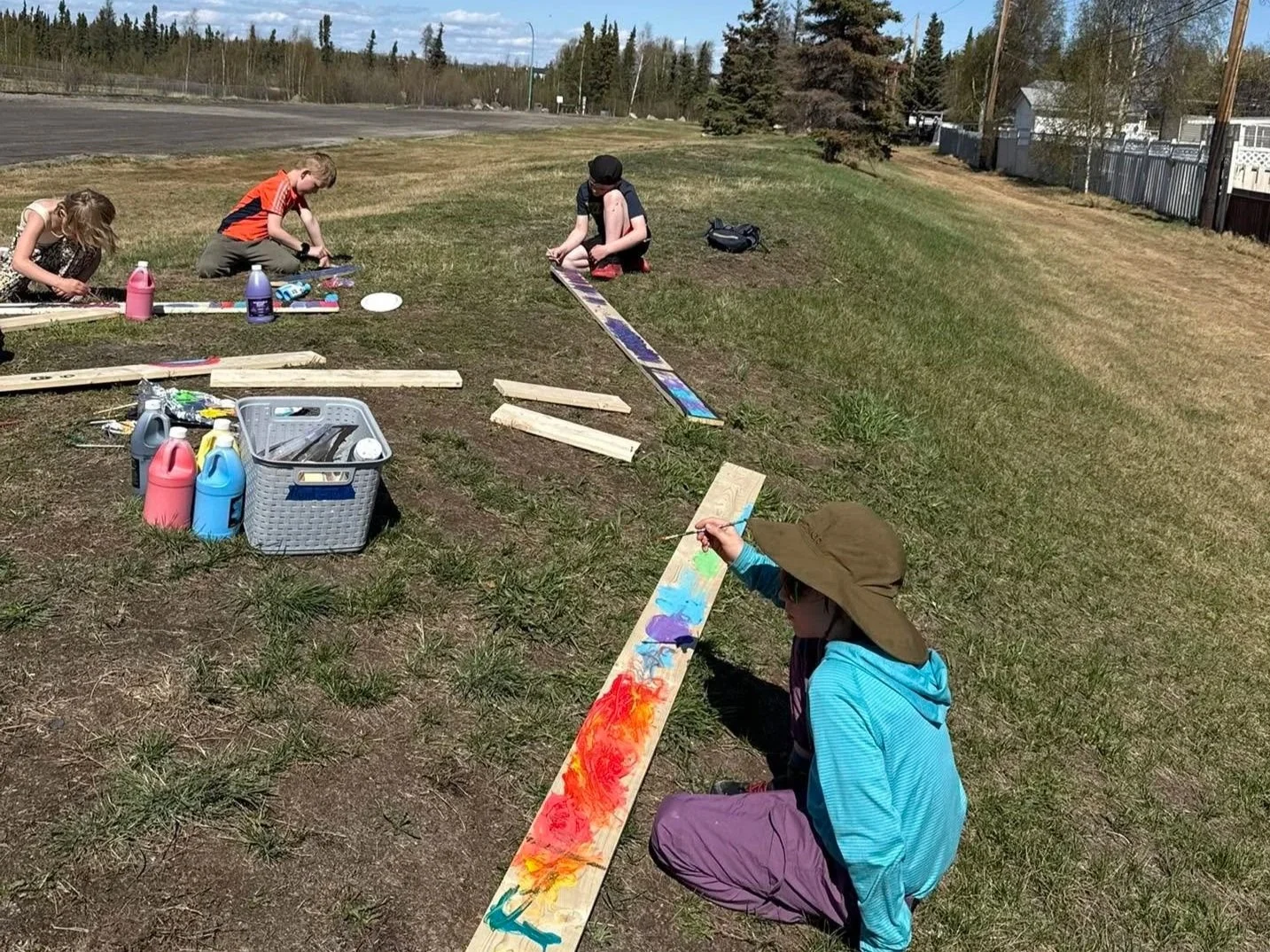 Children painting outdoors on wooden planks laid on grass in a park-like area on a sunny day.