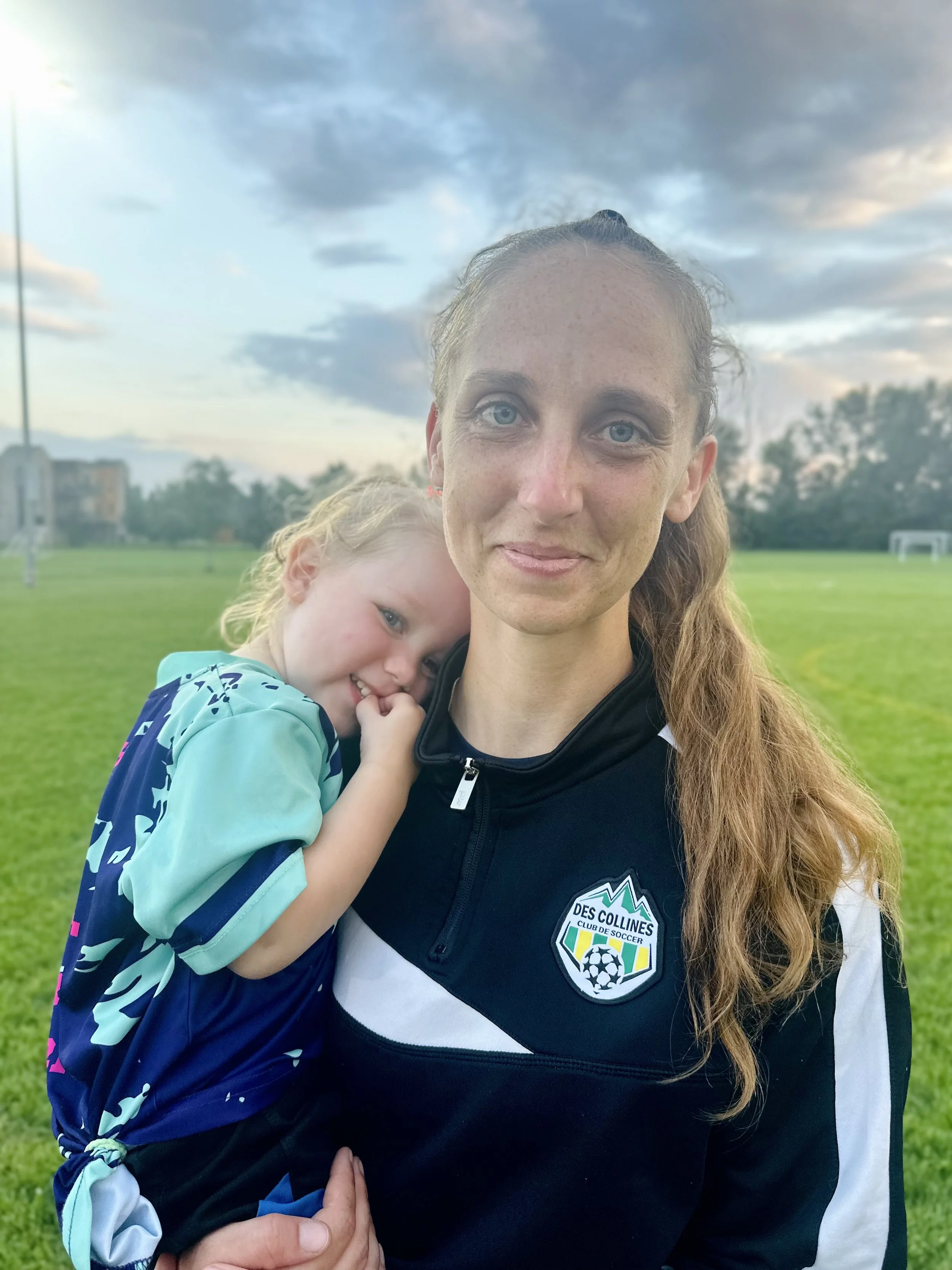 A woman with long, curly hair and a young girl hugging her, standing on a grassy field under a cloudy sky during the daytime. The woman is wearing a soccer jersey and the girl is wearing a blue and black sports jacket.