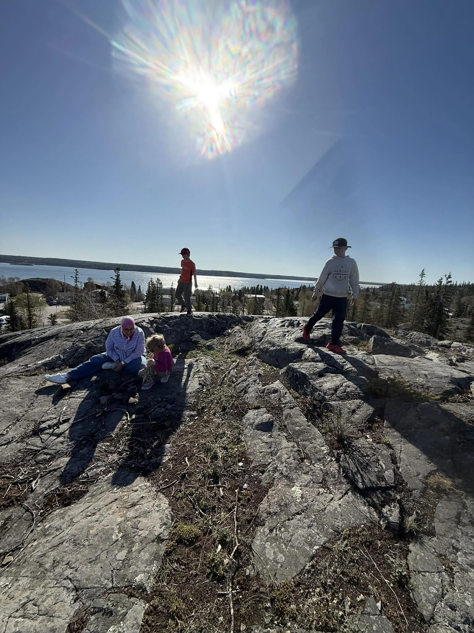 Children and an adult standing and sitting on rocky ground outdoors with a large body of water and trees in the background, under a bright sun with lens flare in the sky.