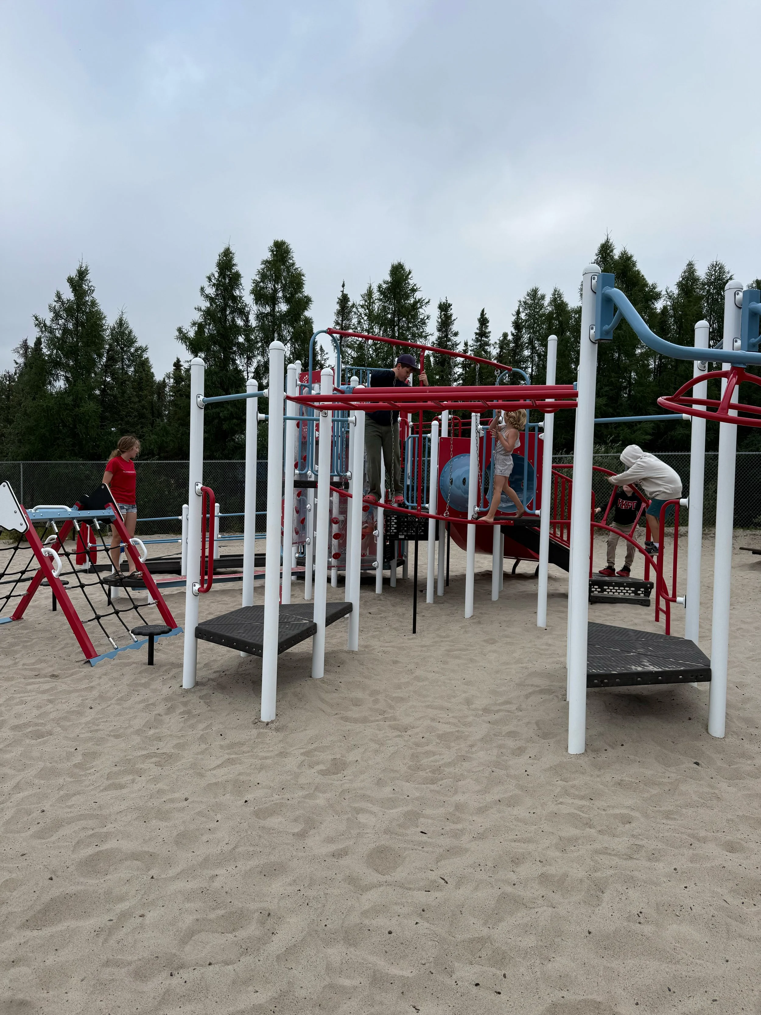 Children and a man playing on a colorful outdoor playground at a sand-covered park, with trees and a cloudy sky in the background.