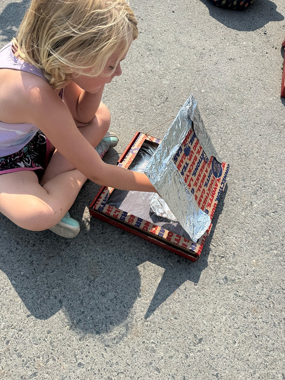 A young girl with blonde hair, wearing a sleeveless top, shorts, and sneakers, crouching on a concrete surface, looking into a cardboard box with an aluminum foil covering on one side, casting a shadow on the ground.