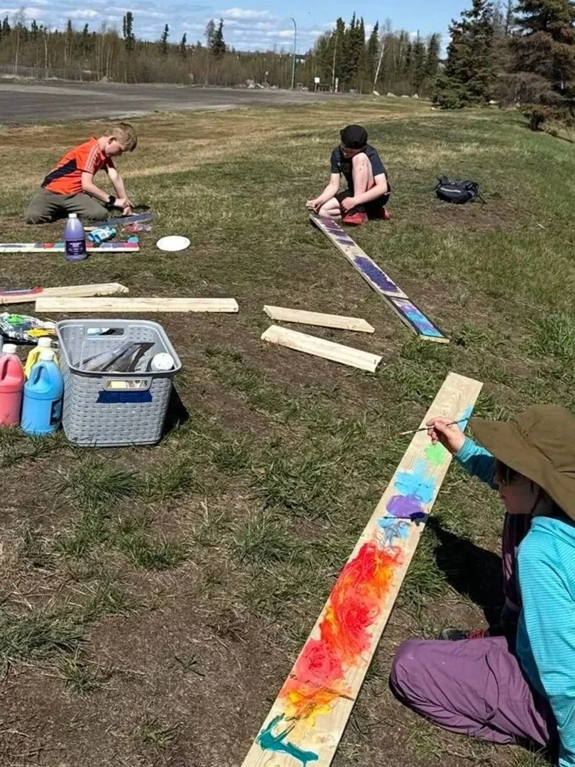 Children painting on wooden planks outdoors on a grassy field with trees and a road in the background.