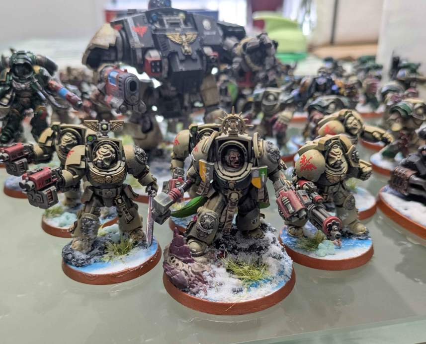 Collection of painted Warhammer 40k miniature figures, including space marines and a large vehicle in the background, all on display with snow and terrain bases.