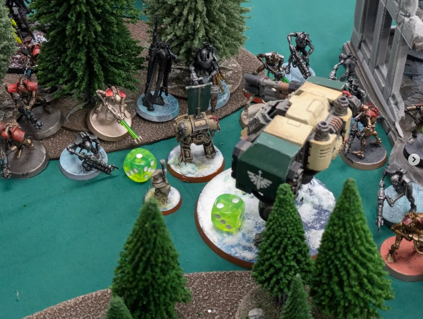 Miniature tabletop war game scene with futuristic soldiers, a large robotic vehicle, trees, and gaming dice on a green surface.
