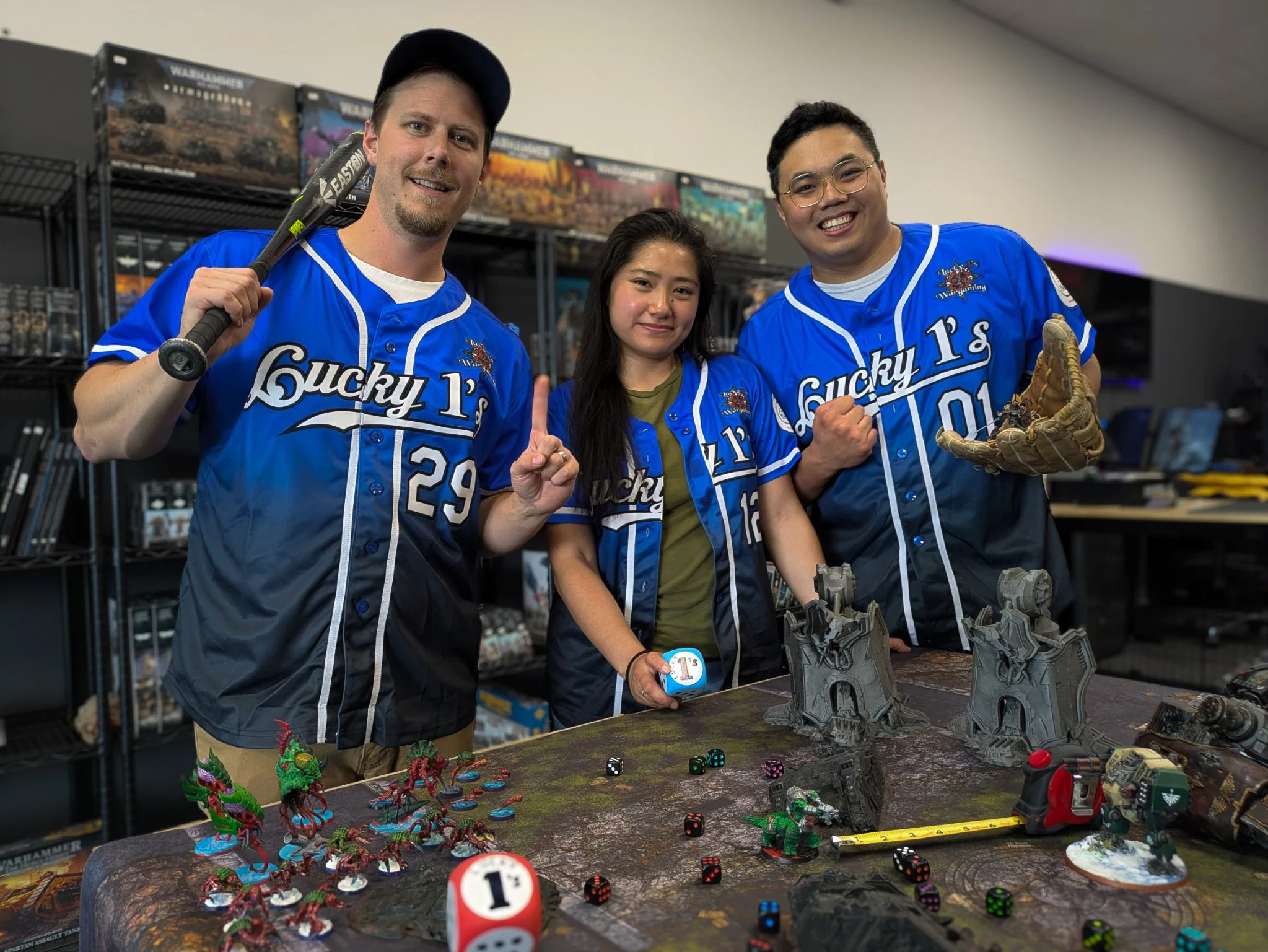 Three people wearing blue sports jerseys with 'Lucky 13' on them are posing at a tabletop gaming event. The table features miniature figurines of monsters and dice. The background has shelves with games.