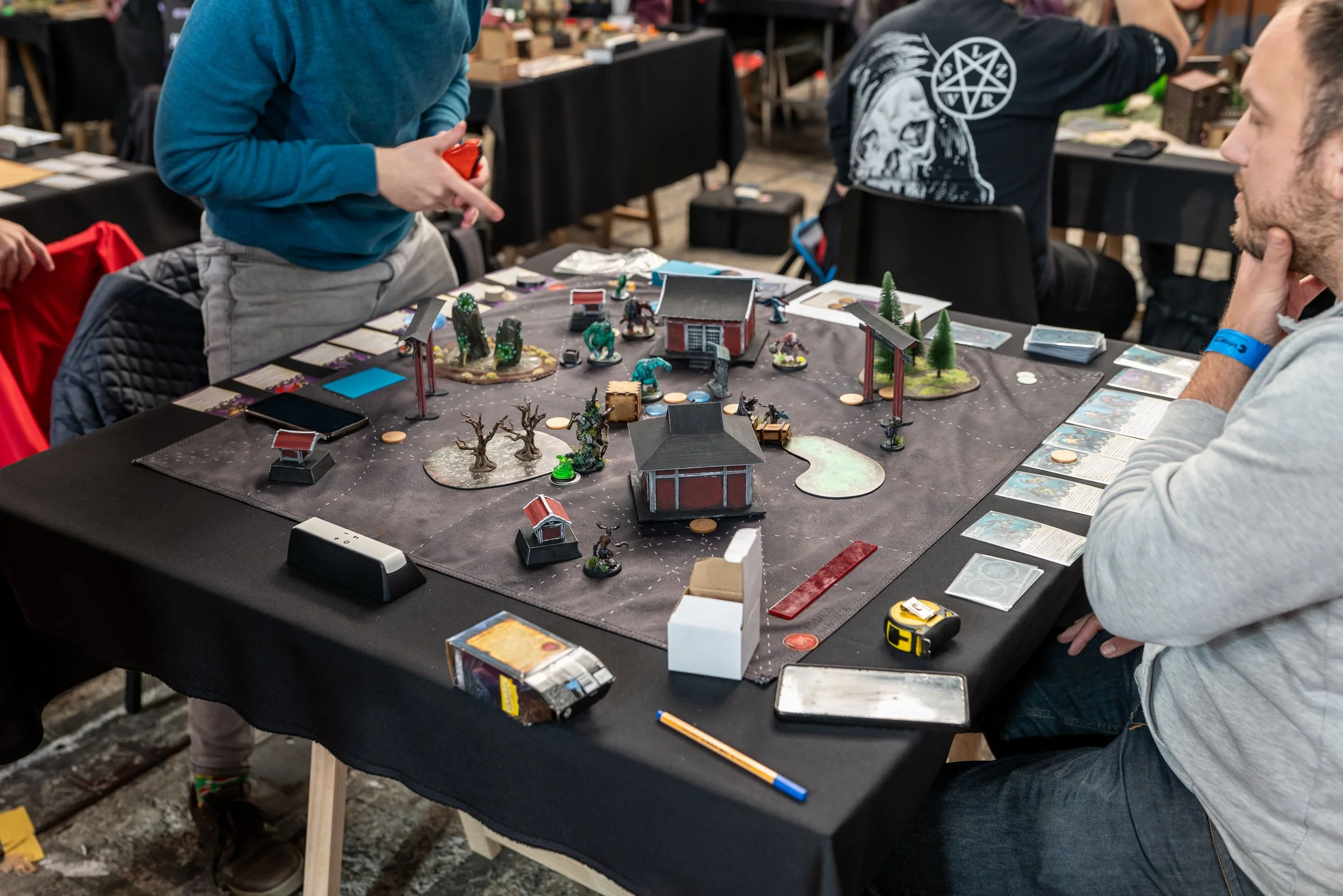 Table with tabletop game pieces, miniature models, and rule sheets, surrounded by players at a gaming event.